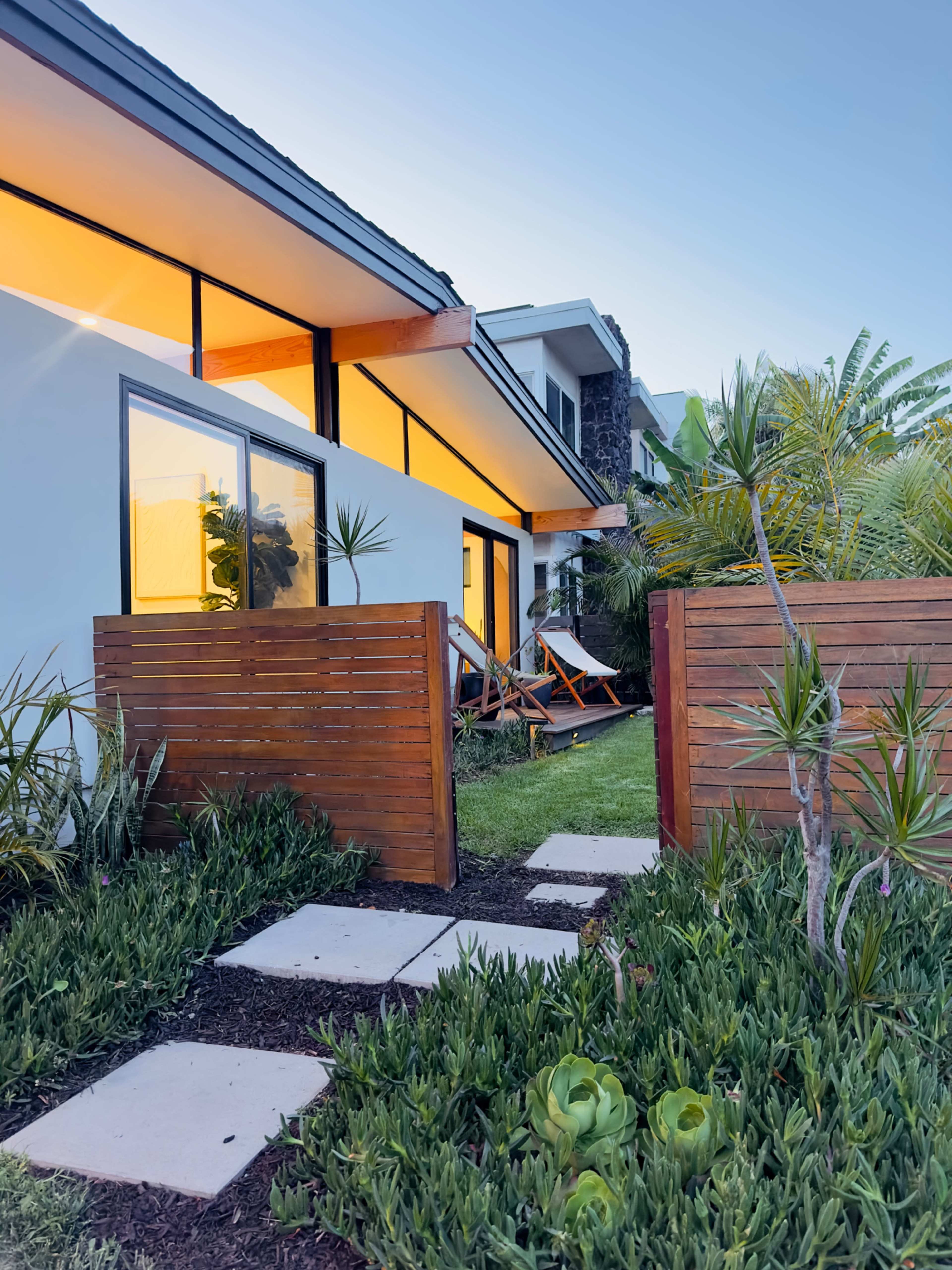 The image shows a modern house with large windows and a landscaped garden featuring a pathway made of stone tiles and wooden fence sections.
