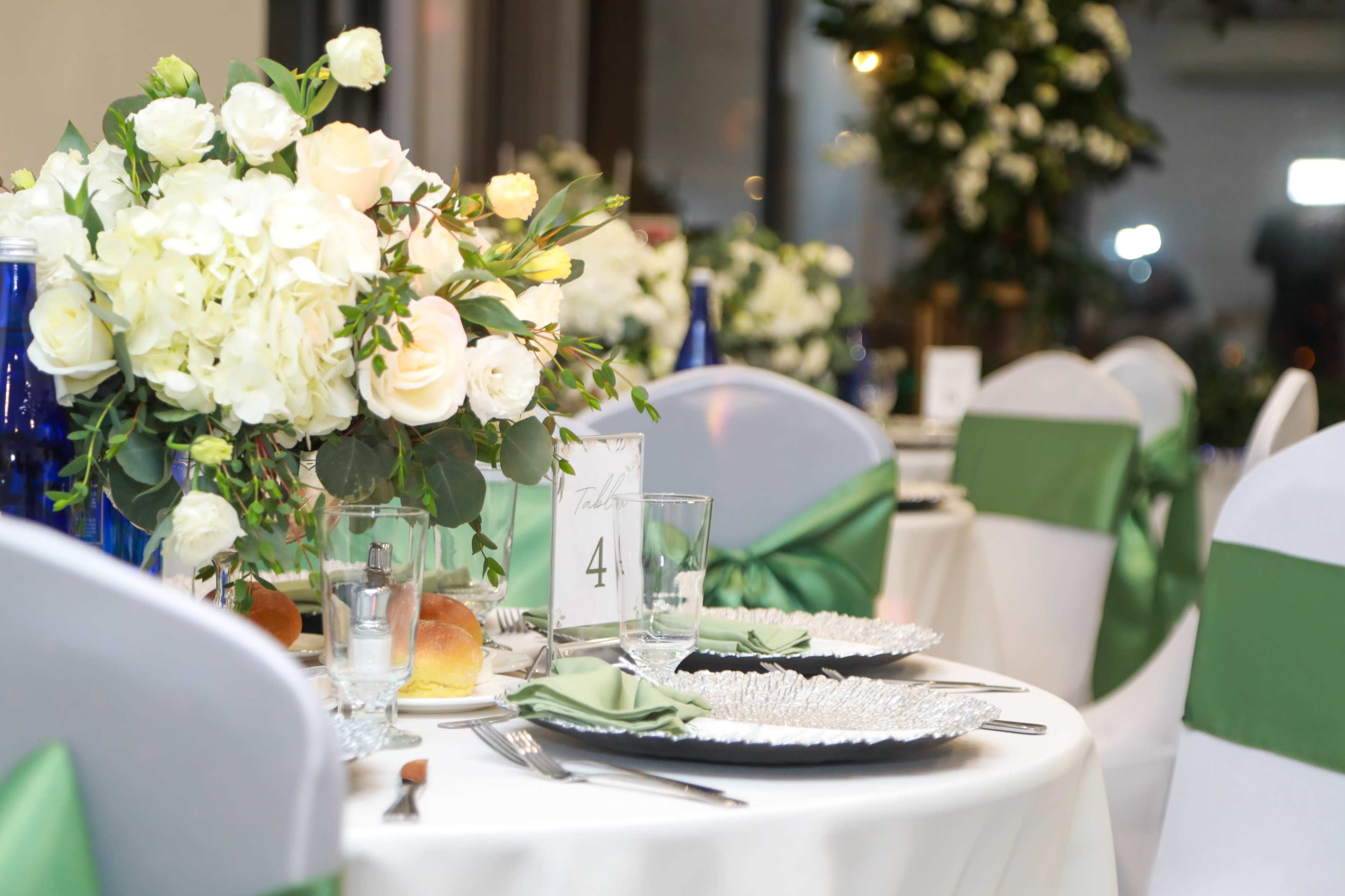 The image shows a elegantly set dining table adorned with white floral arrangements, silver plates, and green decorations in a well-lit venue.