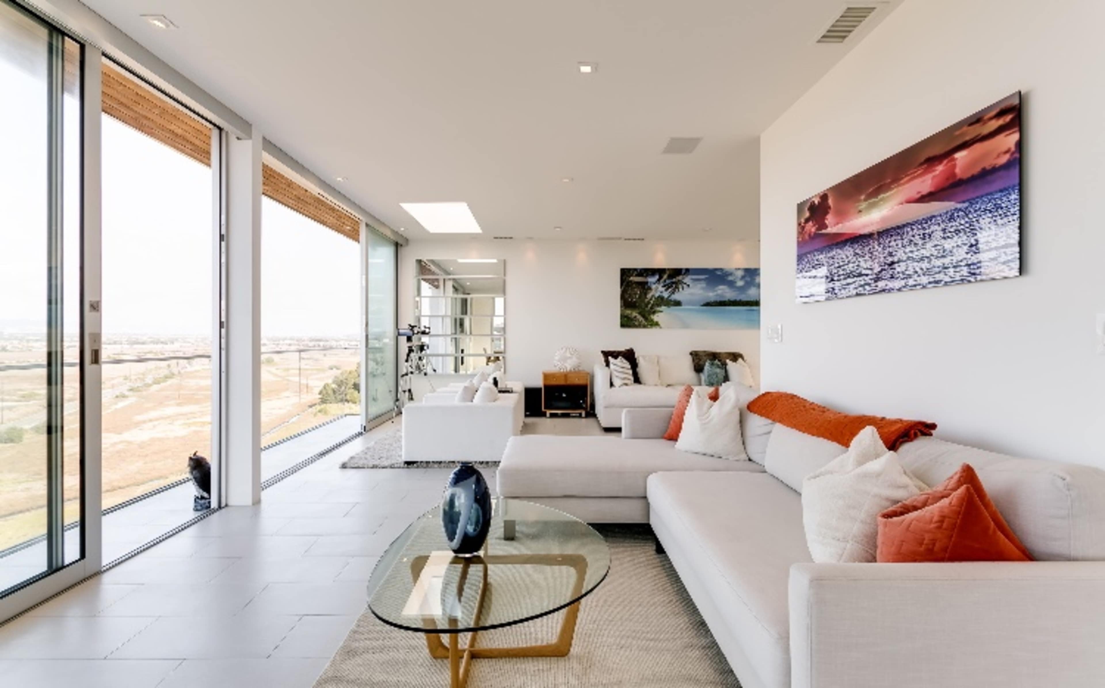 A modern living room with large glass windows overlooking a landscape, featuring light-colored sofas, a round glass coffee table, and minimal decor.