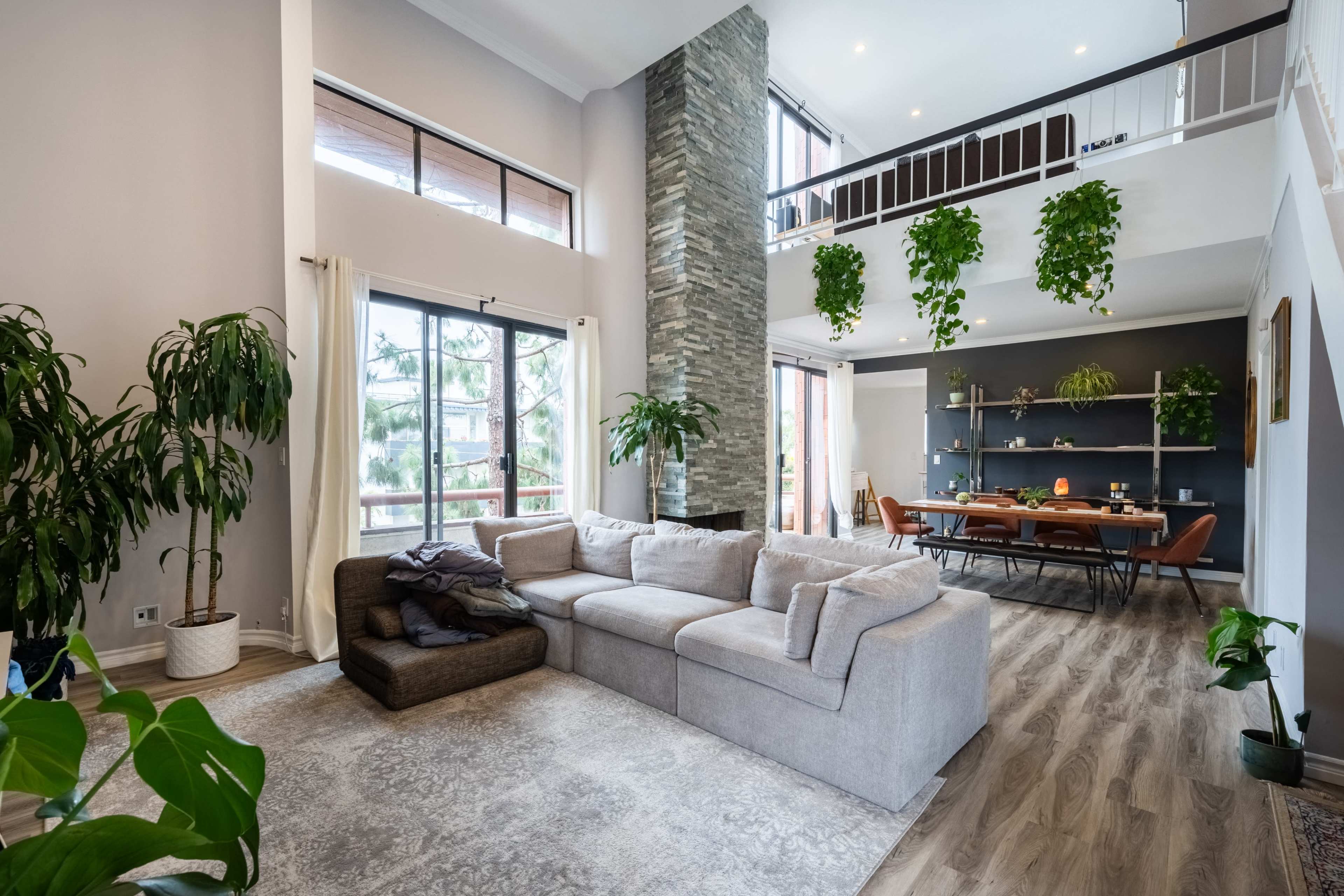A spacious living room with a large sectional sofa, tall windows, and a dining area with a wooden table and plants hanging from the ceiling.