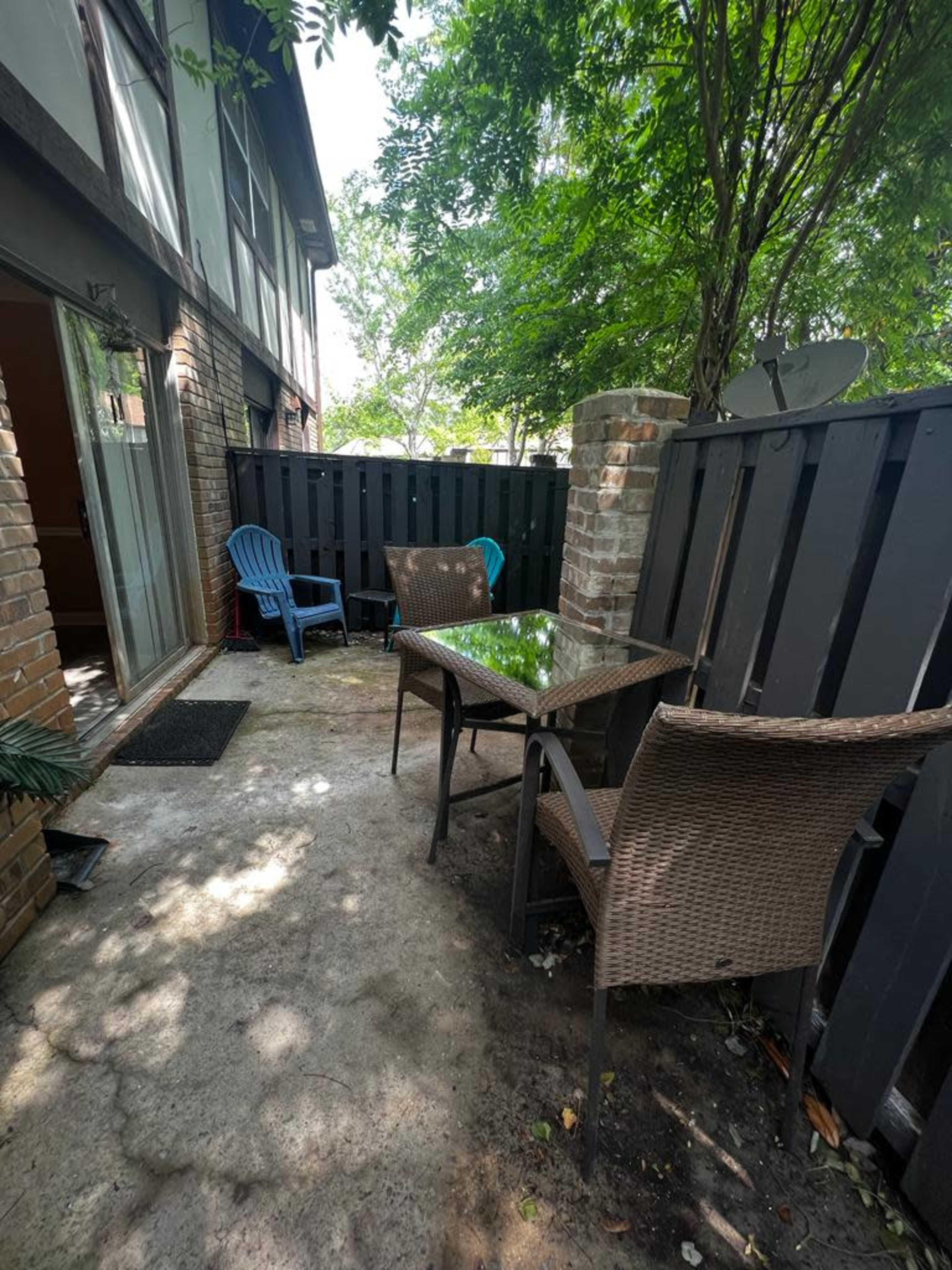 A small patio area features a round glass table surrounded by two wicker chairs, with a blue chair in the corner and greenery overhead.