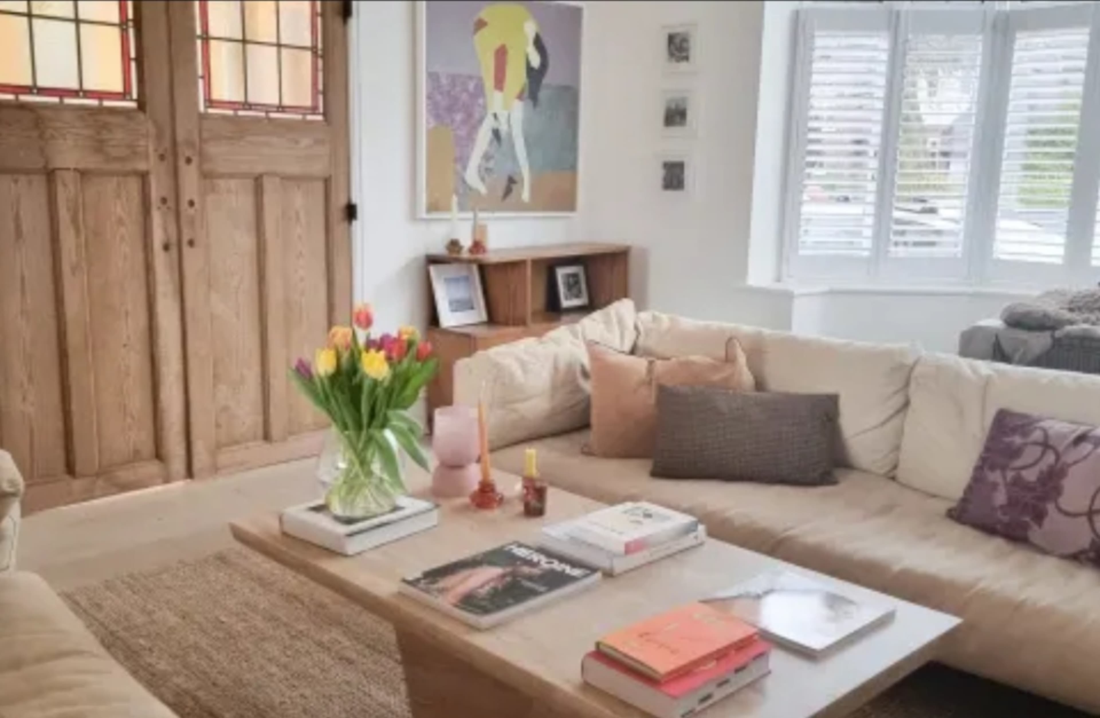 A living room features a beige sectional sofa, a wooden coffee table with decor items, and a bouquet of tulips on the table, complemented by light-colored walls and large wooden doors.