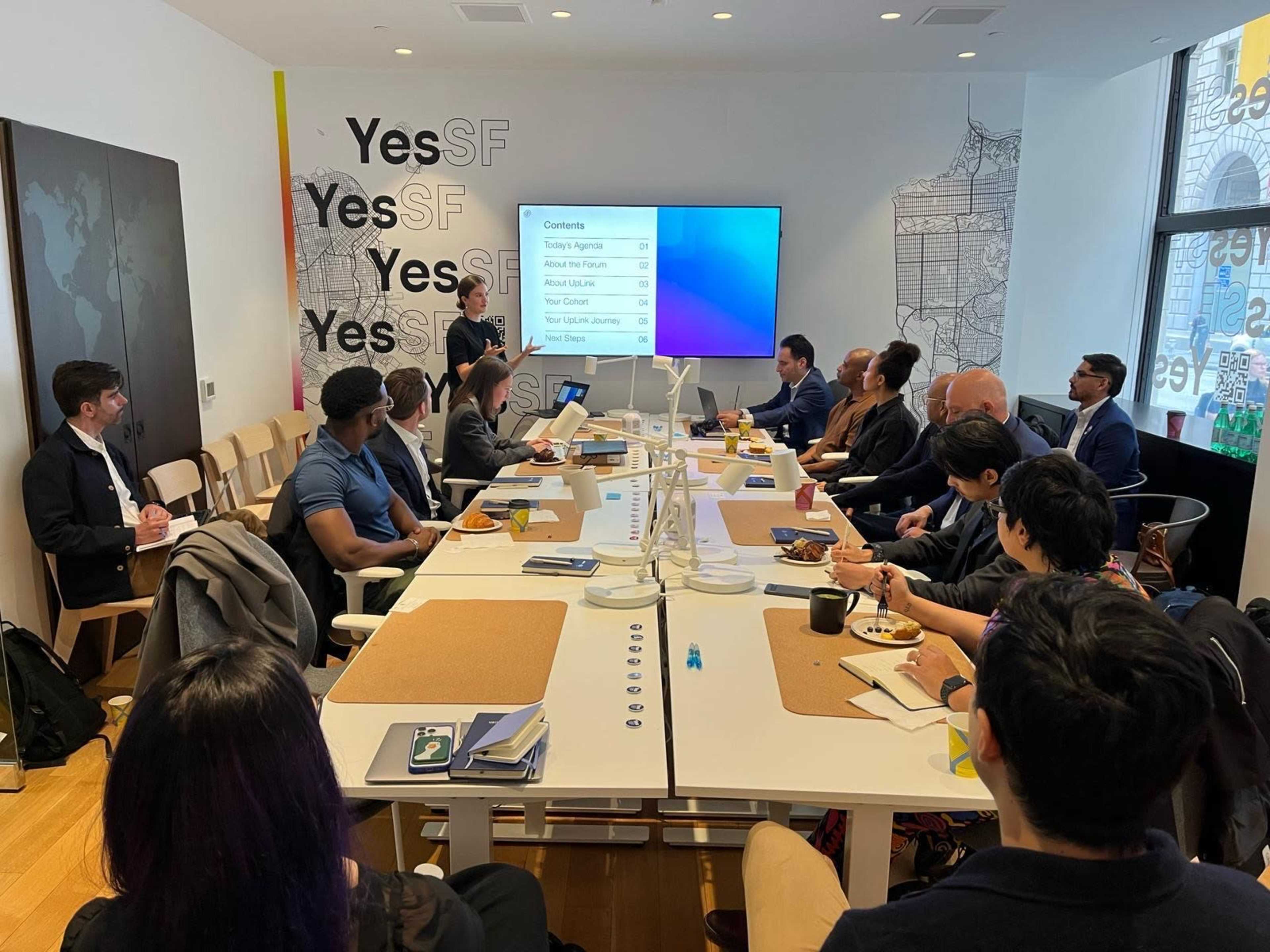 A group of professionals is gathered around a large table in a modern office space, engaging in a presentation displayed on a screen in front of them.