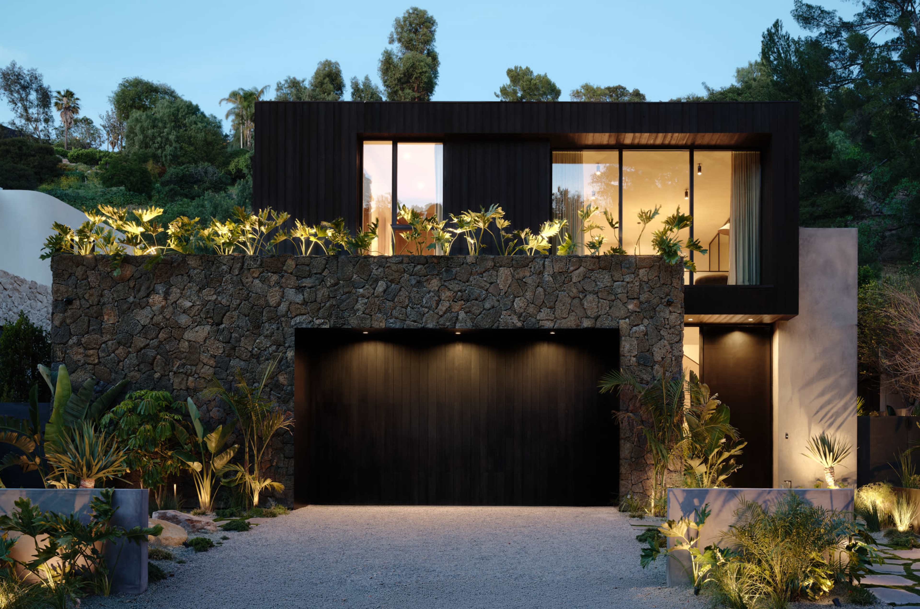 A modern house with a stone facade, large windows, and landscaping features is set against a backdrop of trees.