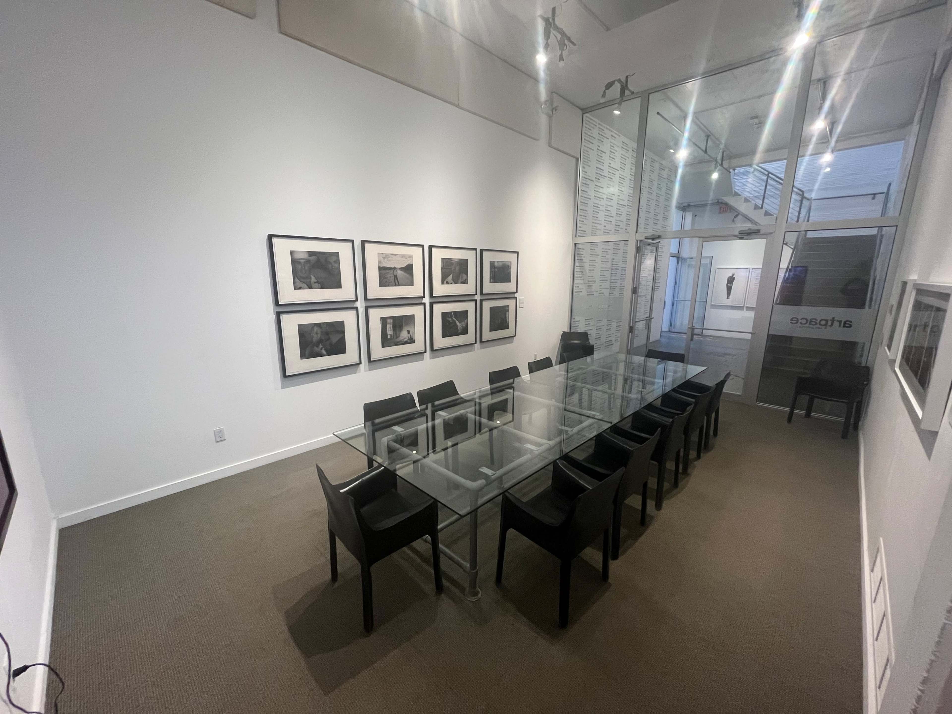 A conference room features a long glass table surrounded by black chairs, with framed photographs on the walls and an entrance on one side.