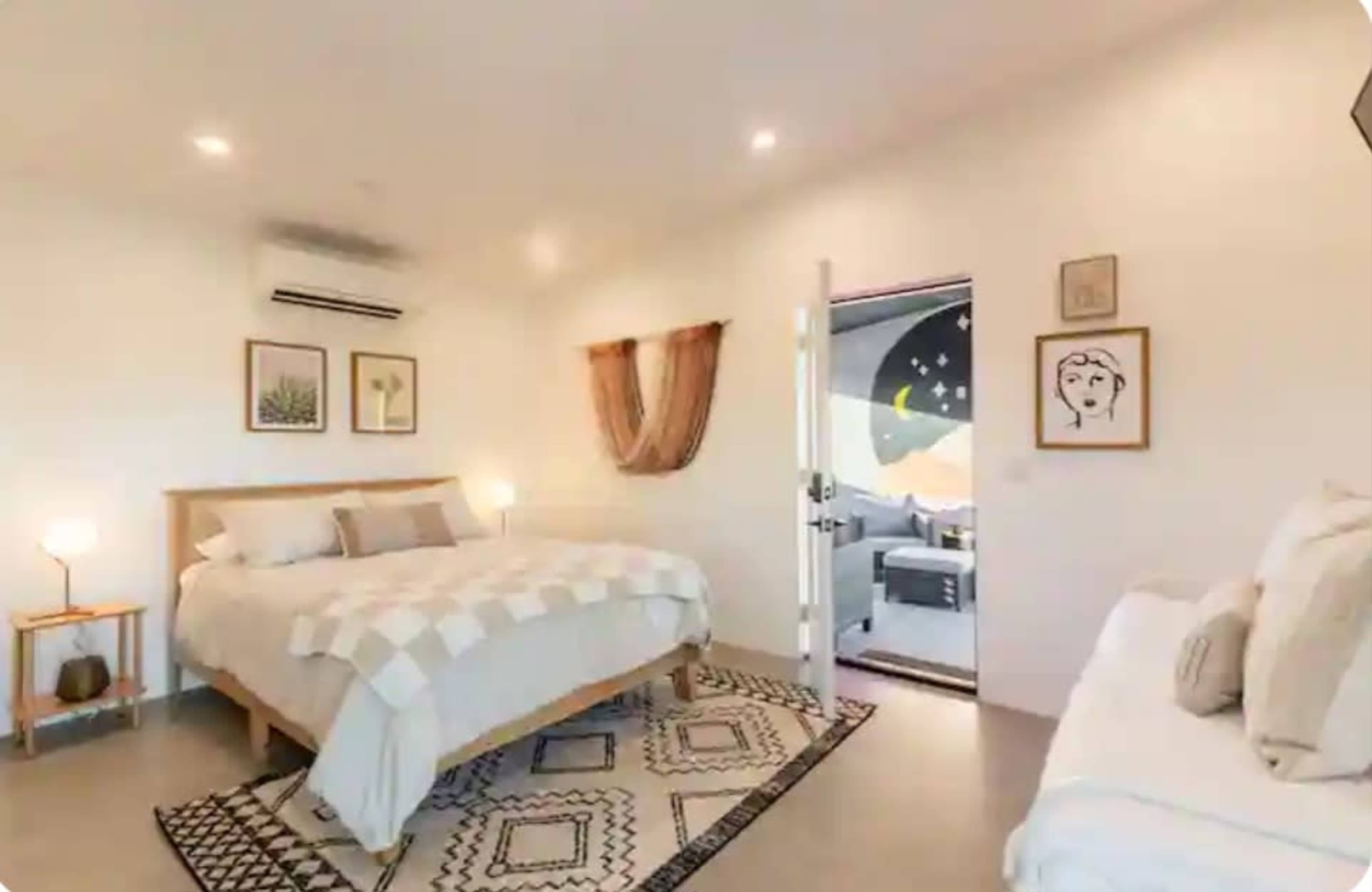 The image shows a well-lit bedroom with a large bed, a smaller bed, and an open door leading to a room with decorative wall art.