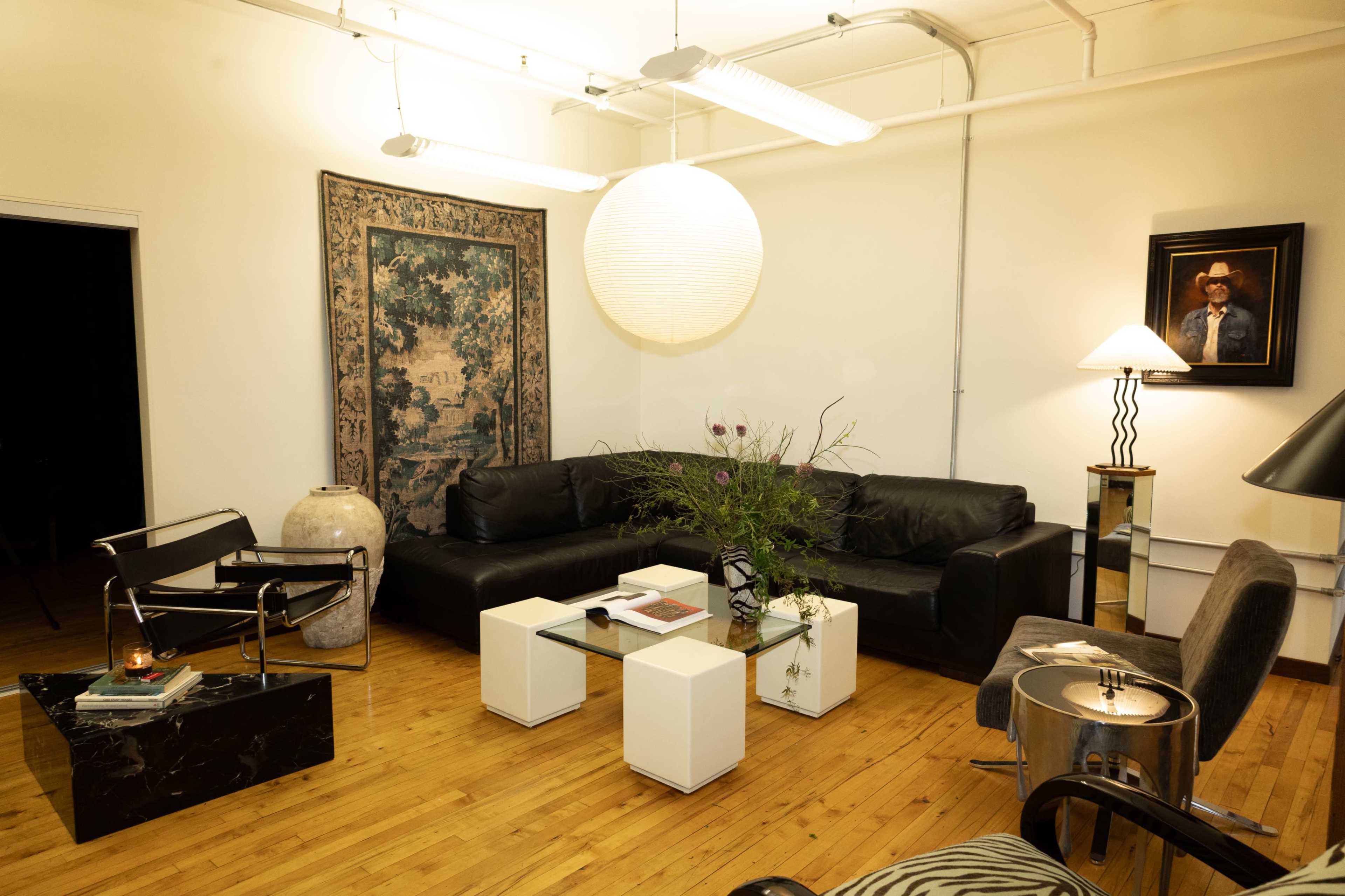 The image shows a modern living space featuring a black leather sofa, a glass coffee table, and various decorative items including a large wall tapestry and a lampshade.
