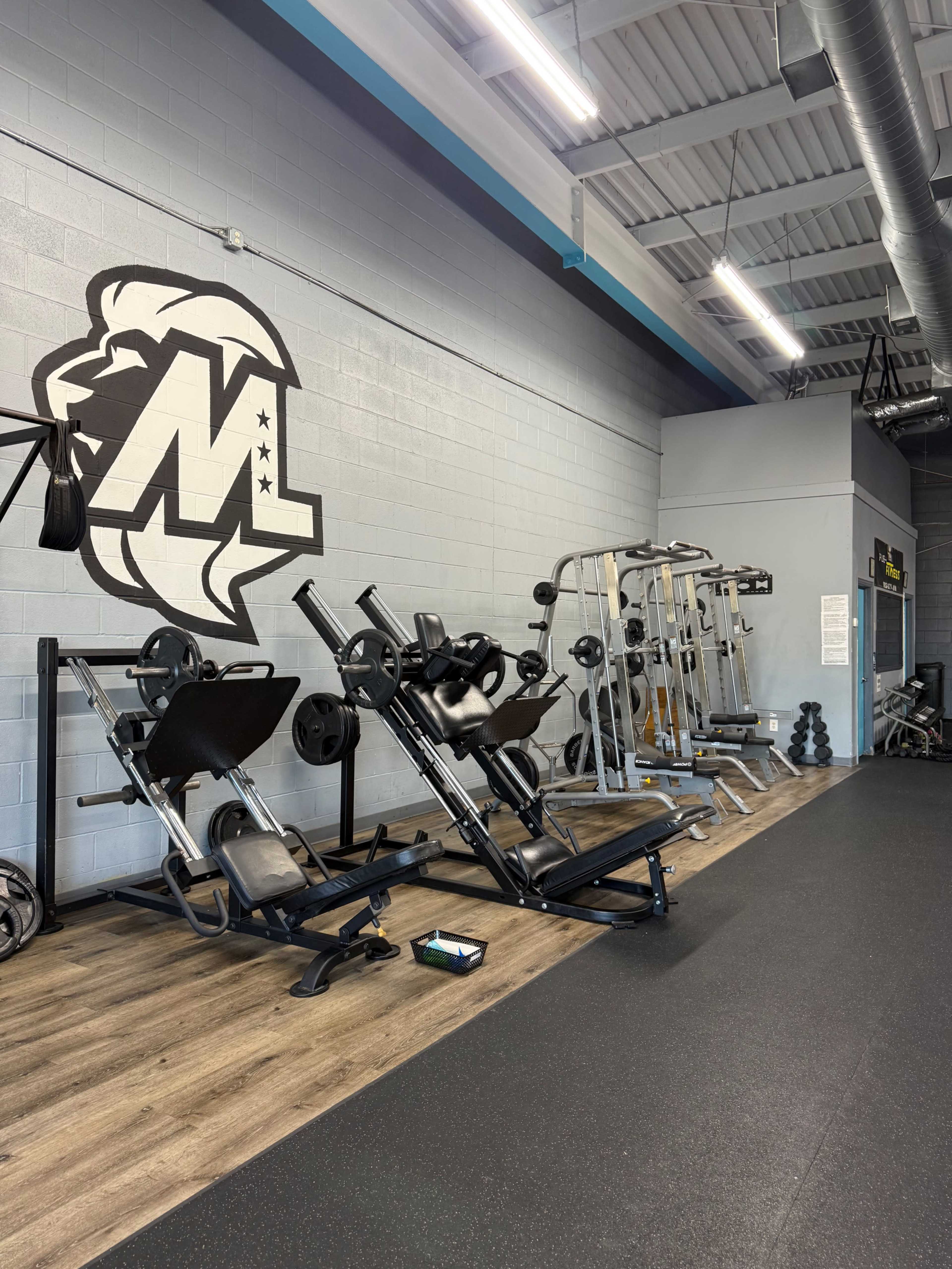 The image shows a gym interior with weightlifting and exercise equipment arranged against a wall featuring a logo.