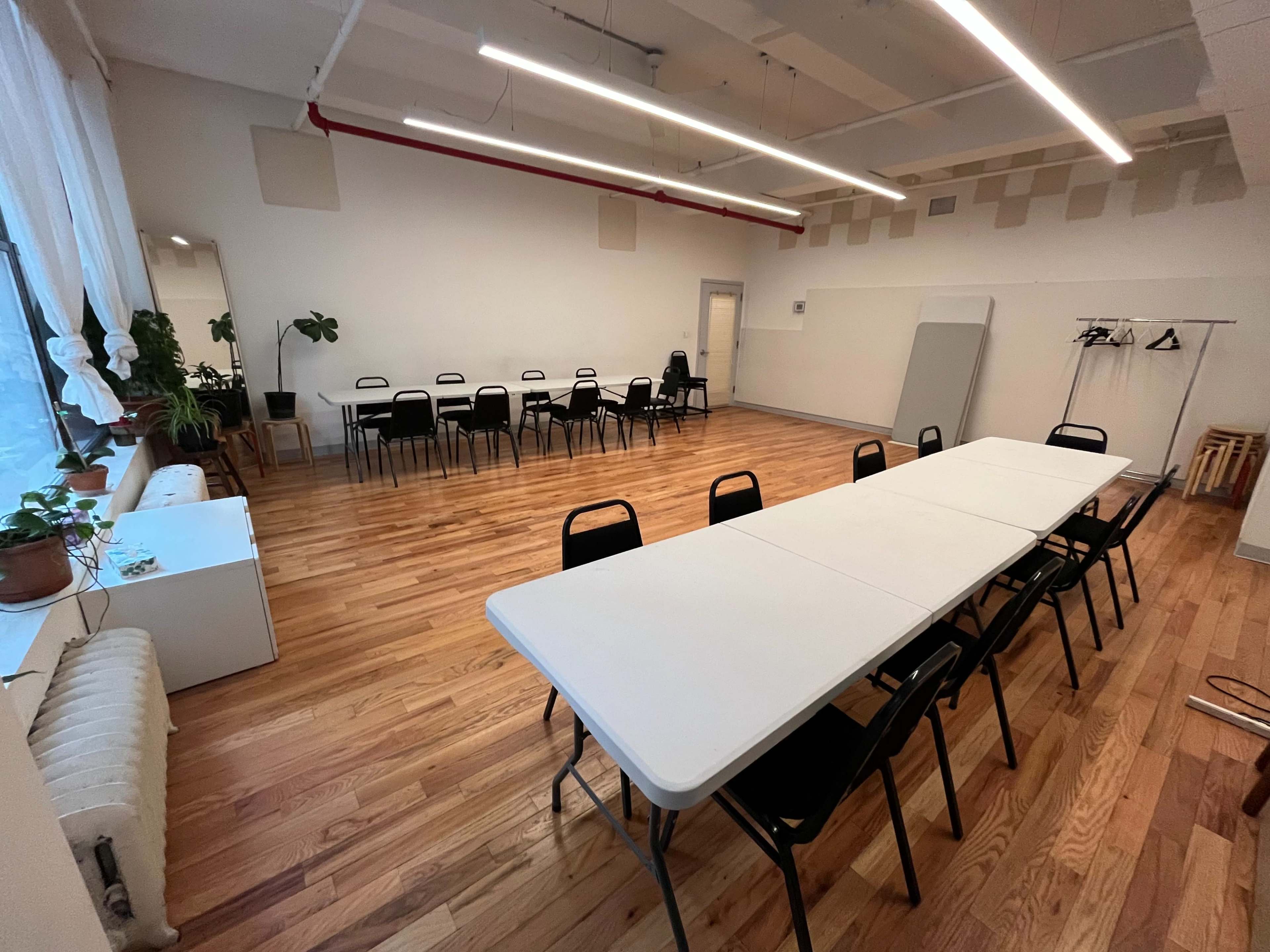 The image shows a spacious room with a long table and chairs arranged for a meeting, featuring hardwood floors and large windows with plants.