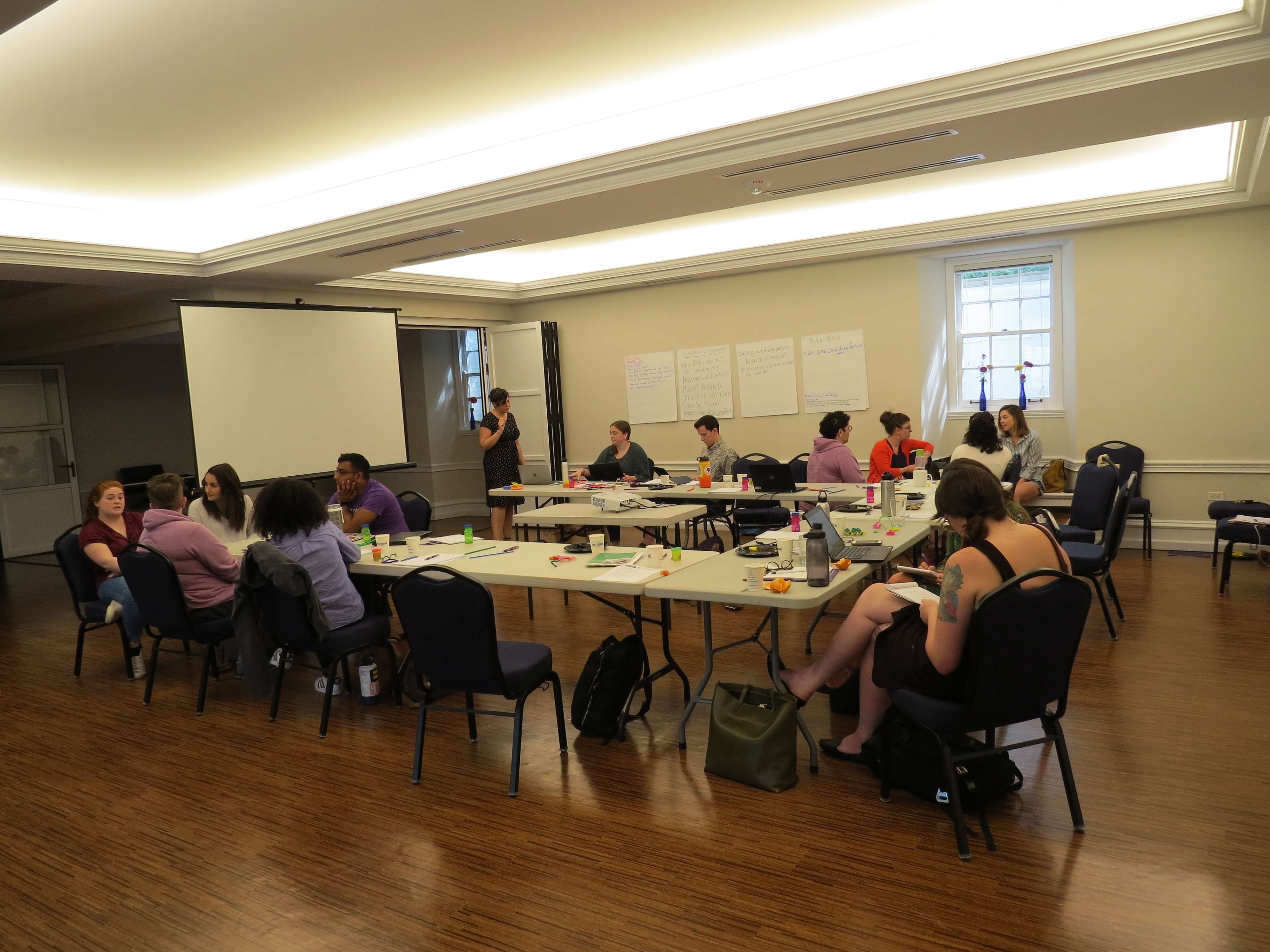 A group of people is seated at tables in a classroom setting, actively engaged in a workshop with a presentation screen and notes displayed on the walls.
