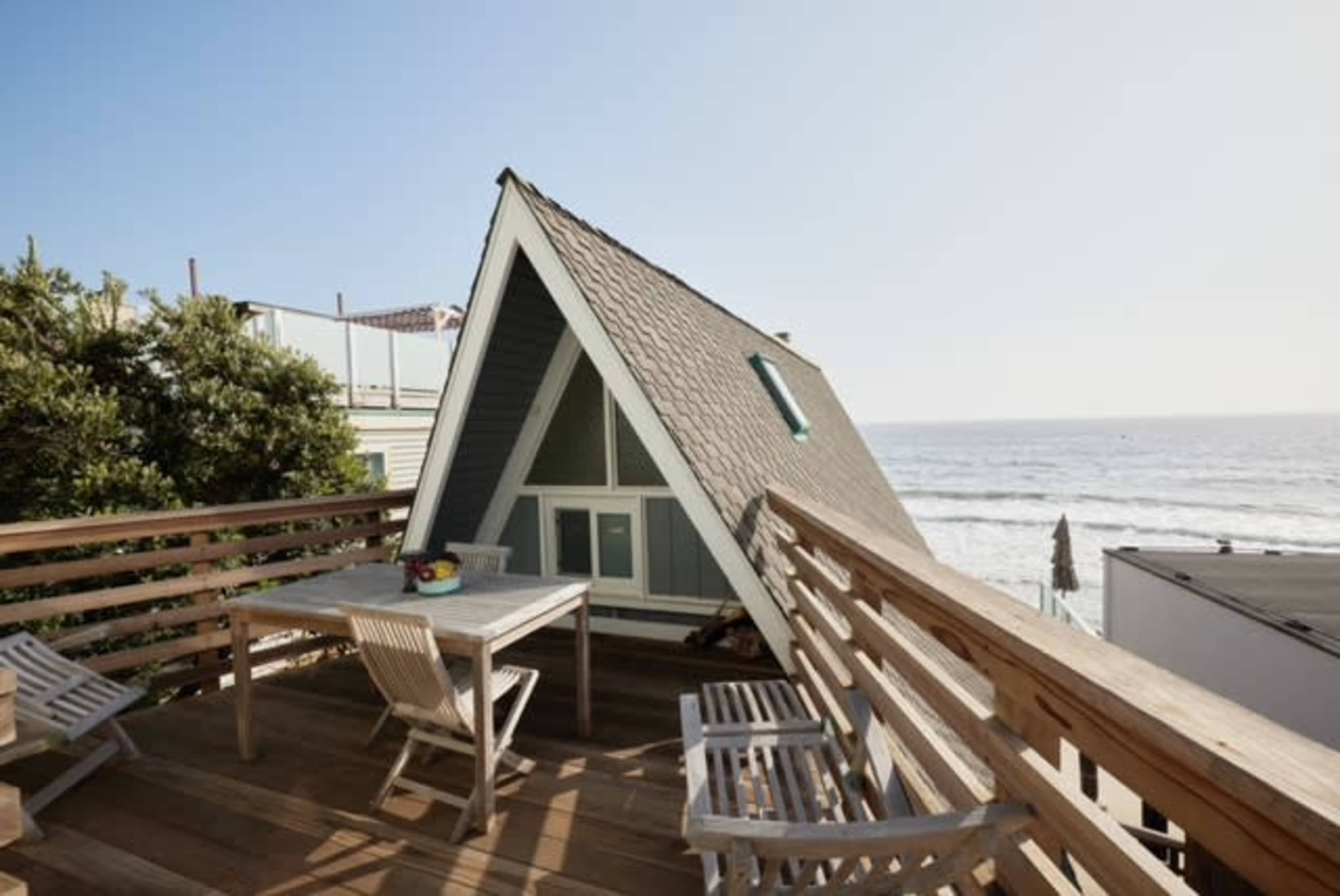 A wooden deck with a table and chairs overlooking a beachside A-frame house and the ocean.