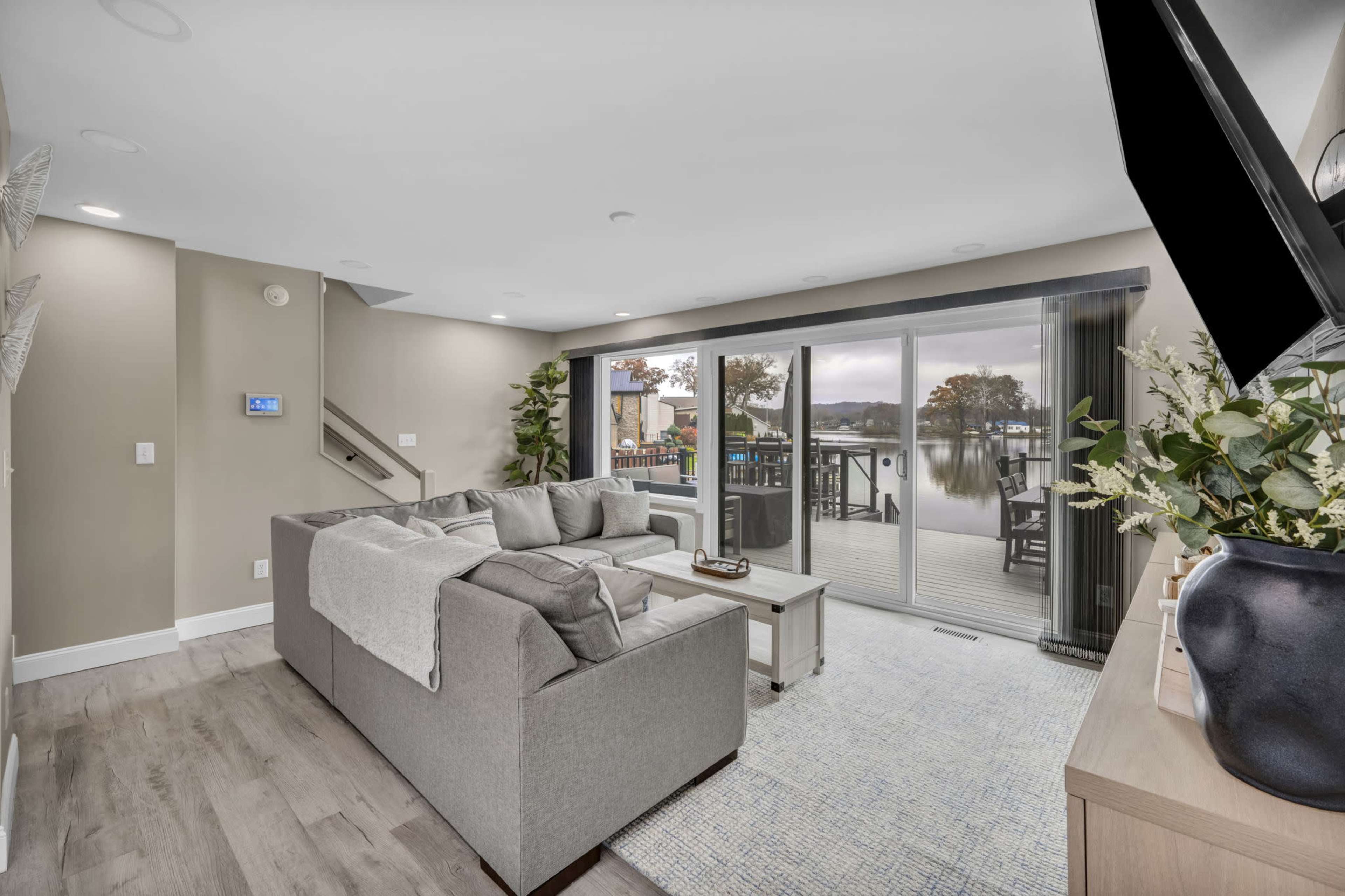The image shows a modern living room with a large gray sofa and a view of a deck by a waterfront through sliding glass doors.