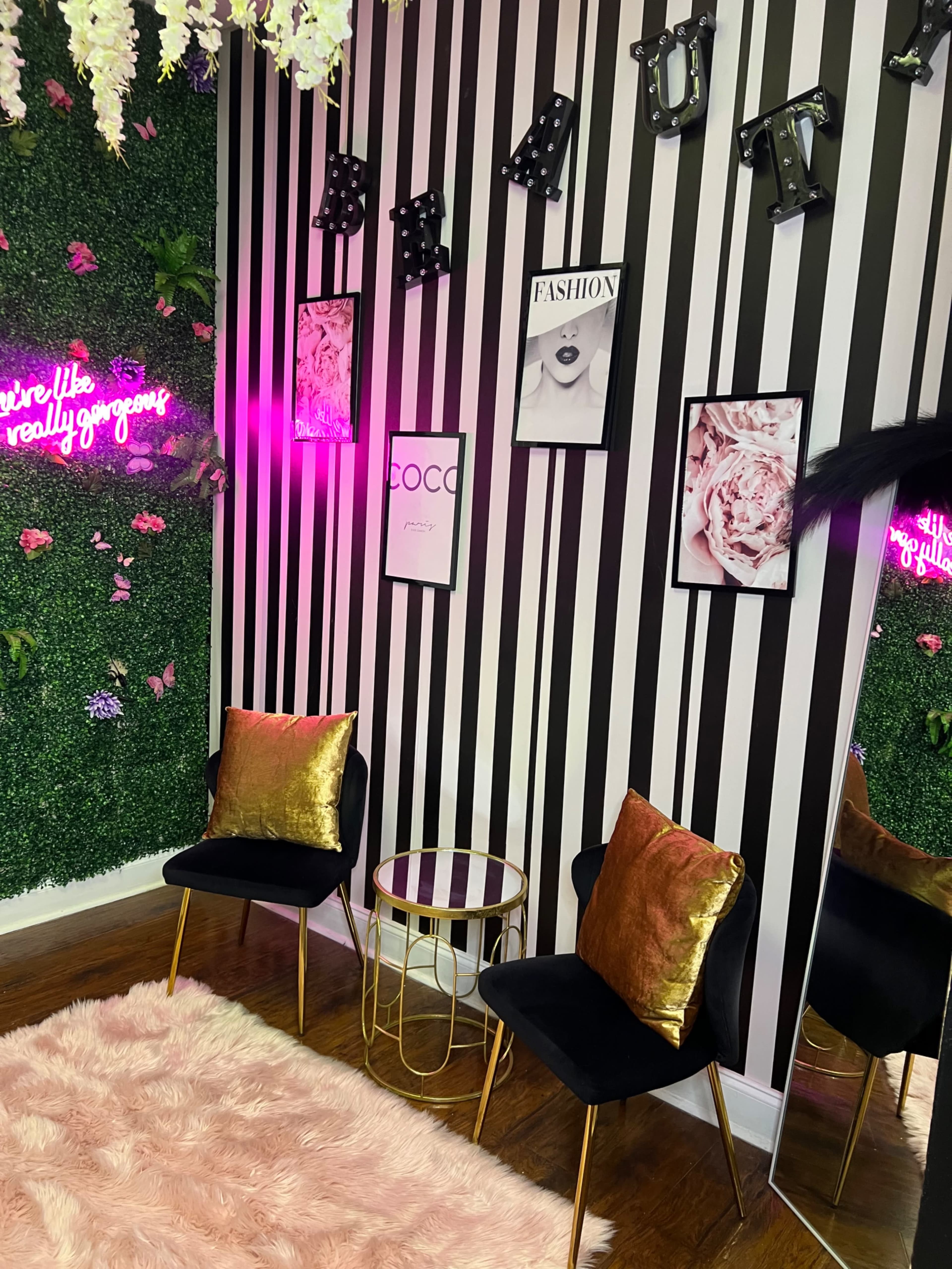 The scene features a stylish corner with striped black and white walls, a faux plant wall, two black chairs with golden pillows, a round table, and framed fashion art.