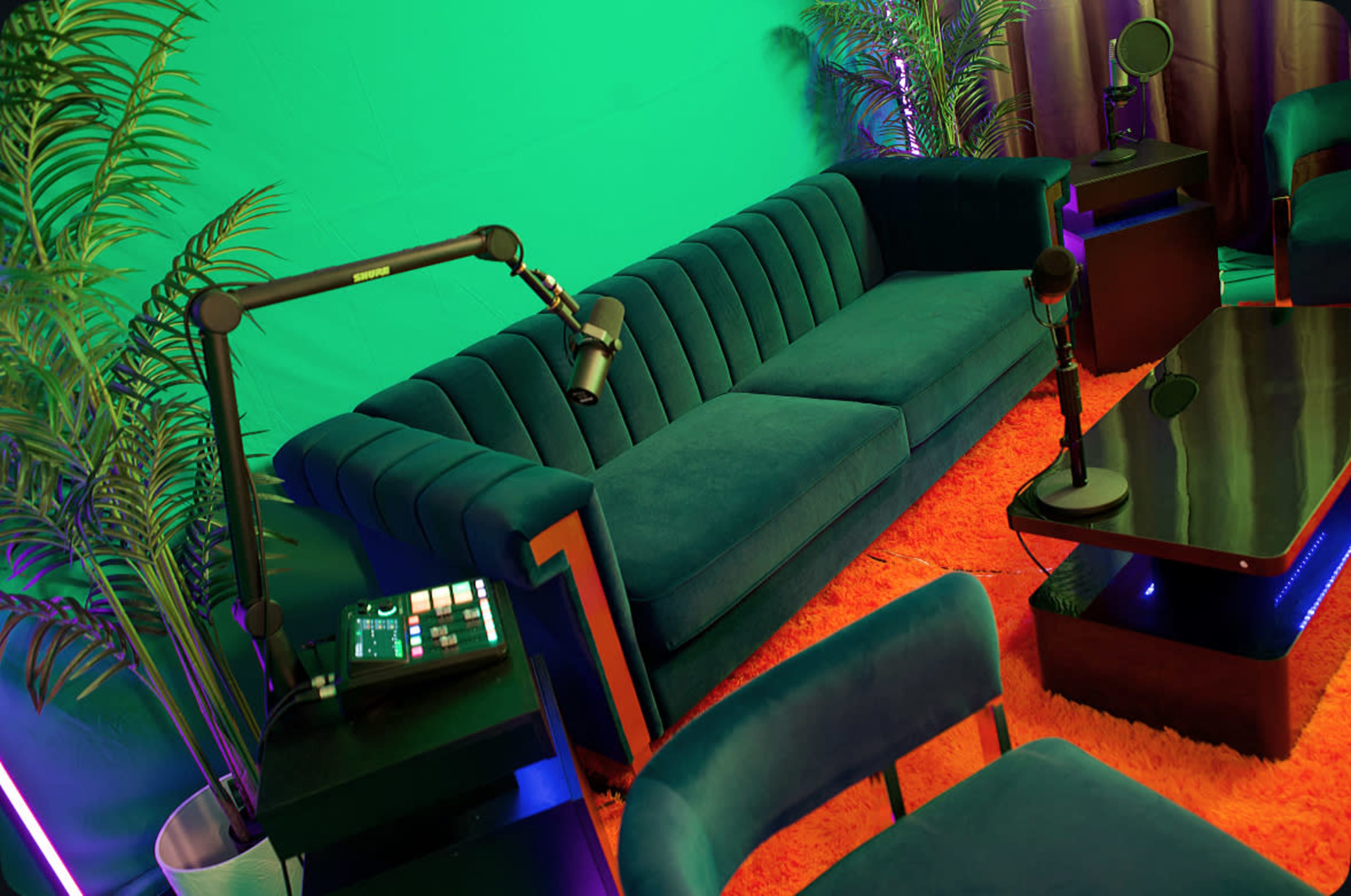 The image shows a modern podcasting studio featuring a plush blue sofa, microphones, and a coffee table, all set against a vibrant green backdrop and illuminated by ambient lighting.