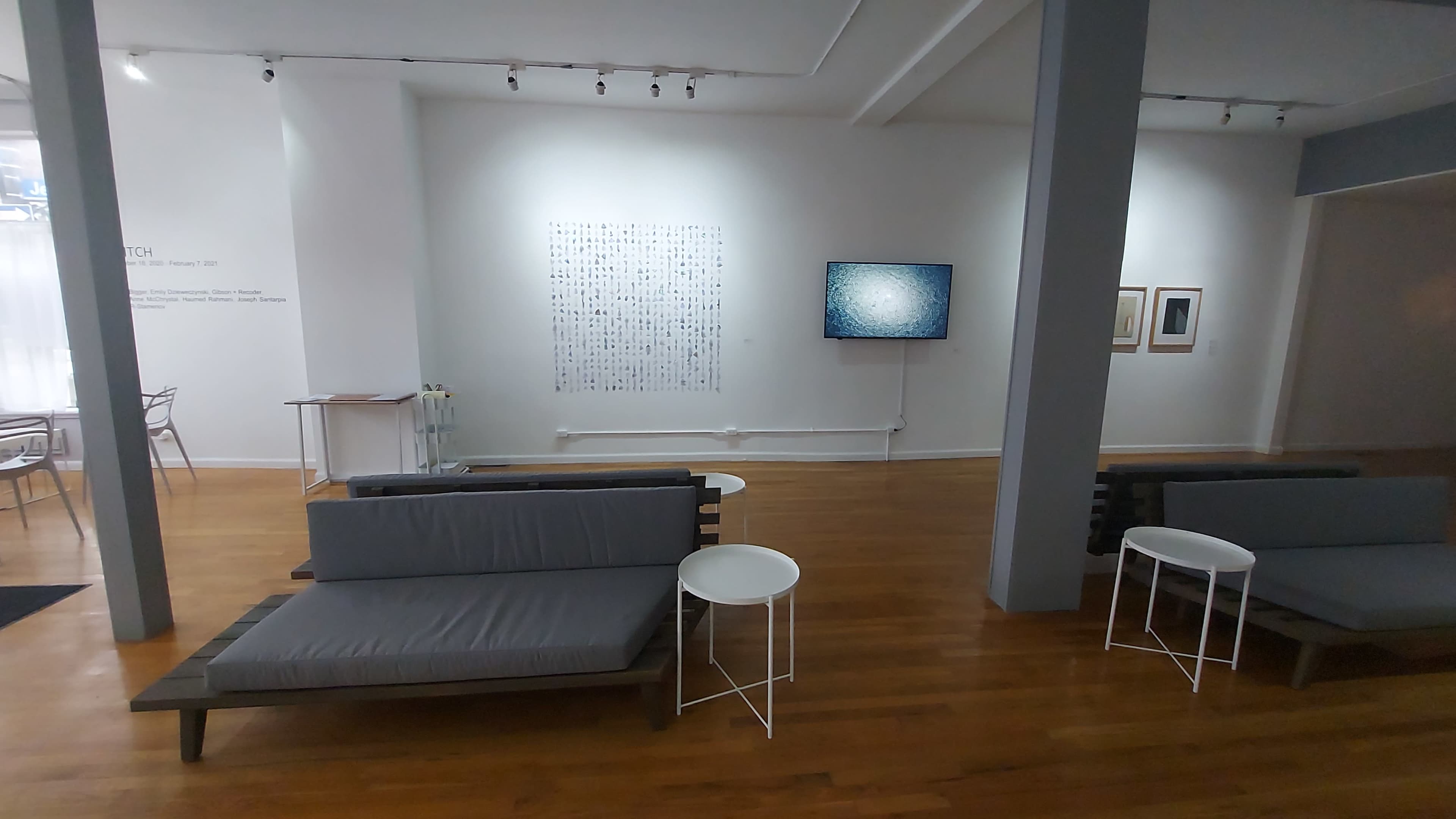 A modern art gallery with minimalistic furniture, including gray couches and white tables, and a wall displaying abstract artwork alongside a television screen.