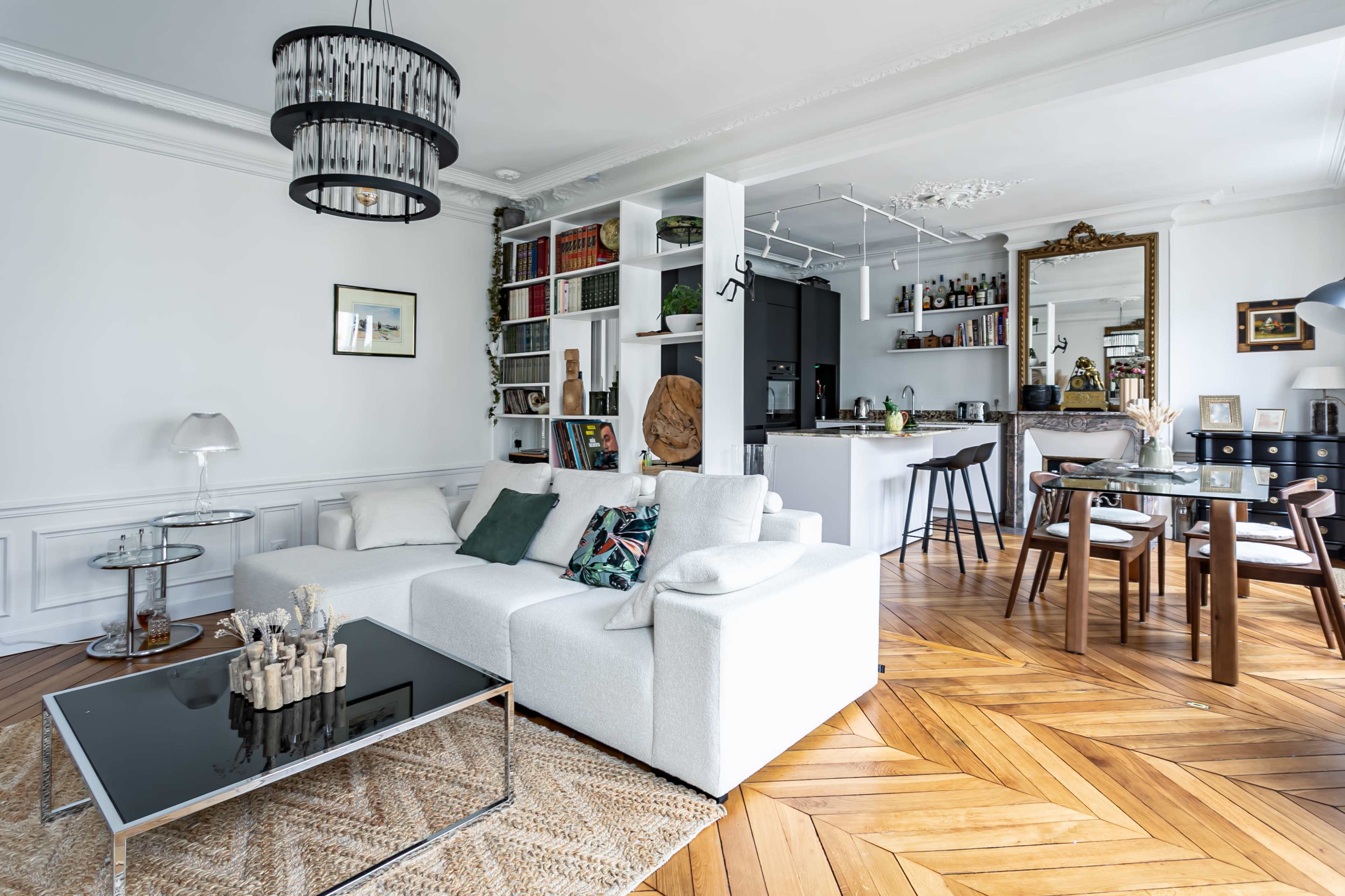 A modern living room with a white sectional sofa, a glass coffee table, and an open kitchen-dining area with stylish decor and wooden flooring.