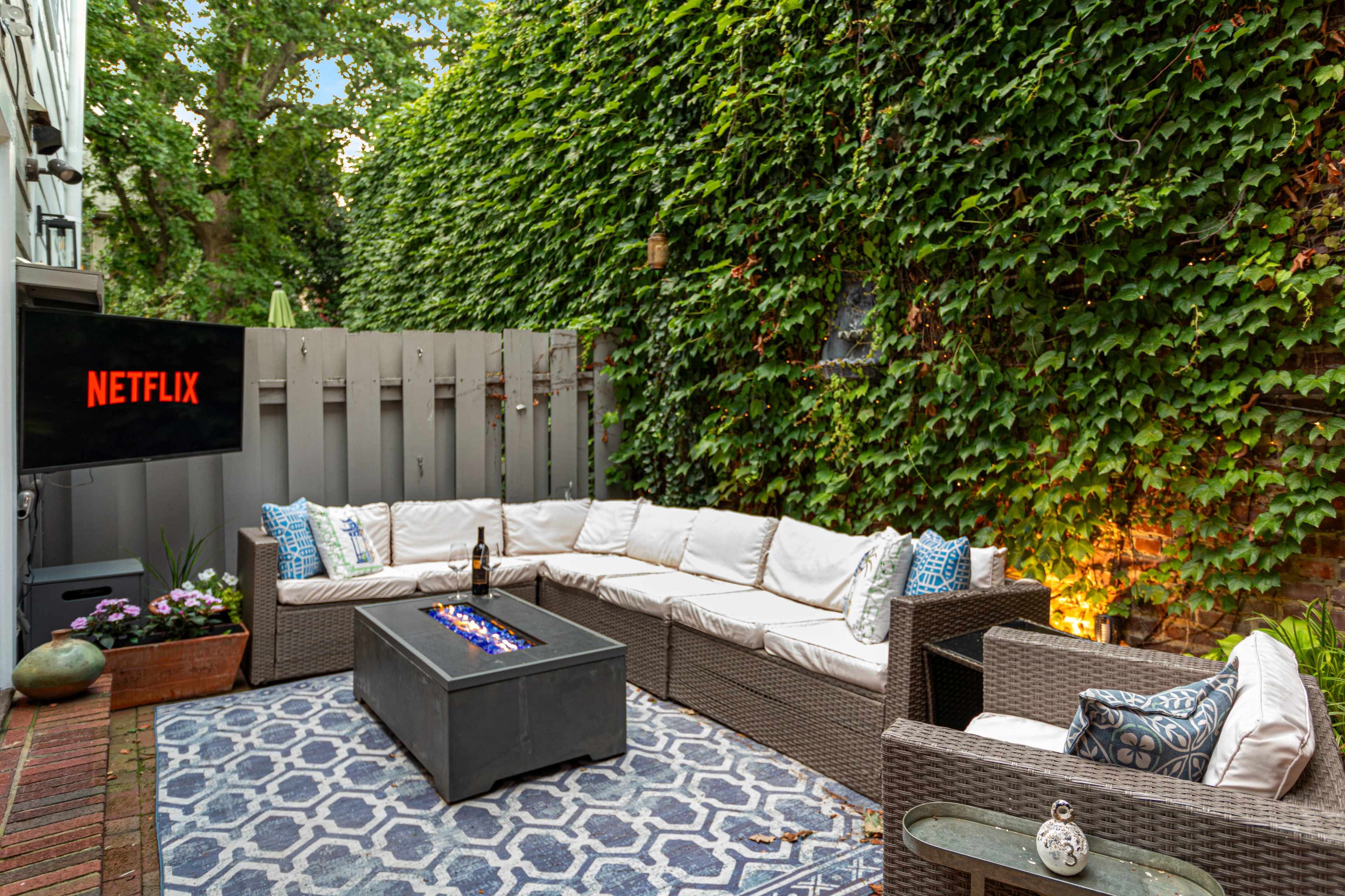 The image features a cozy outdoor seating area with a sectional couch, a fire pit, and a television displaying the Netflix logo, surrounded by lush green ivy.