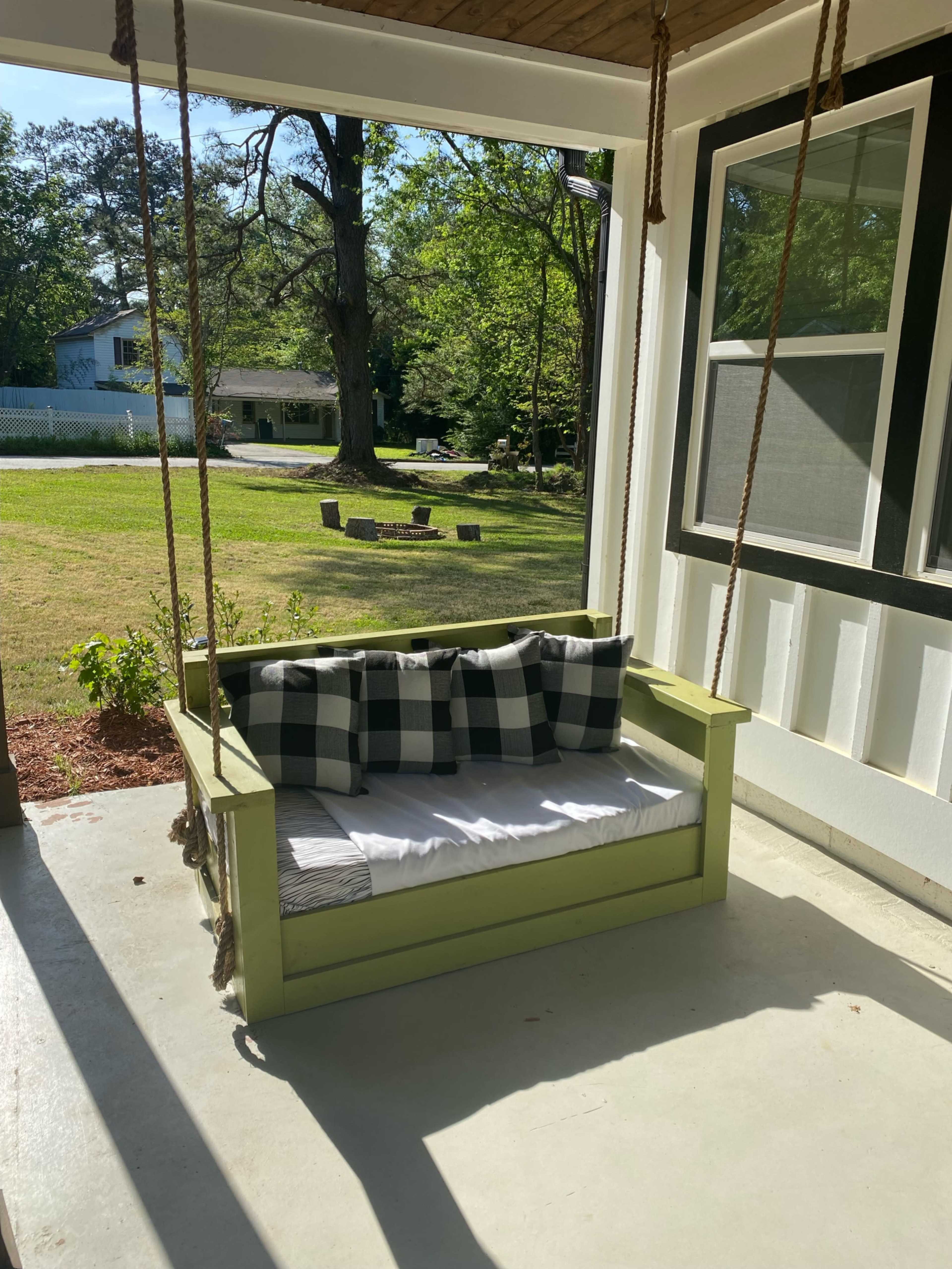A green porch swing with black and white checkered cushions is suspended from the ceiling, overlooking a grassy yard with trees and a house in the background.