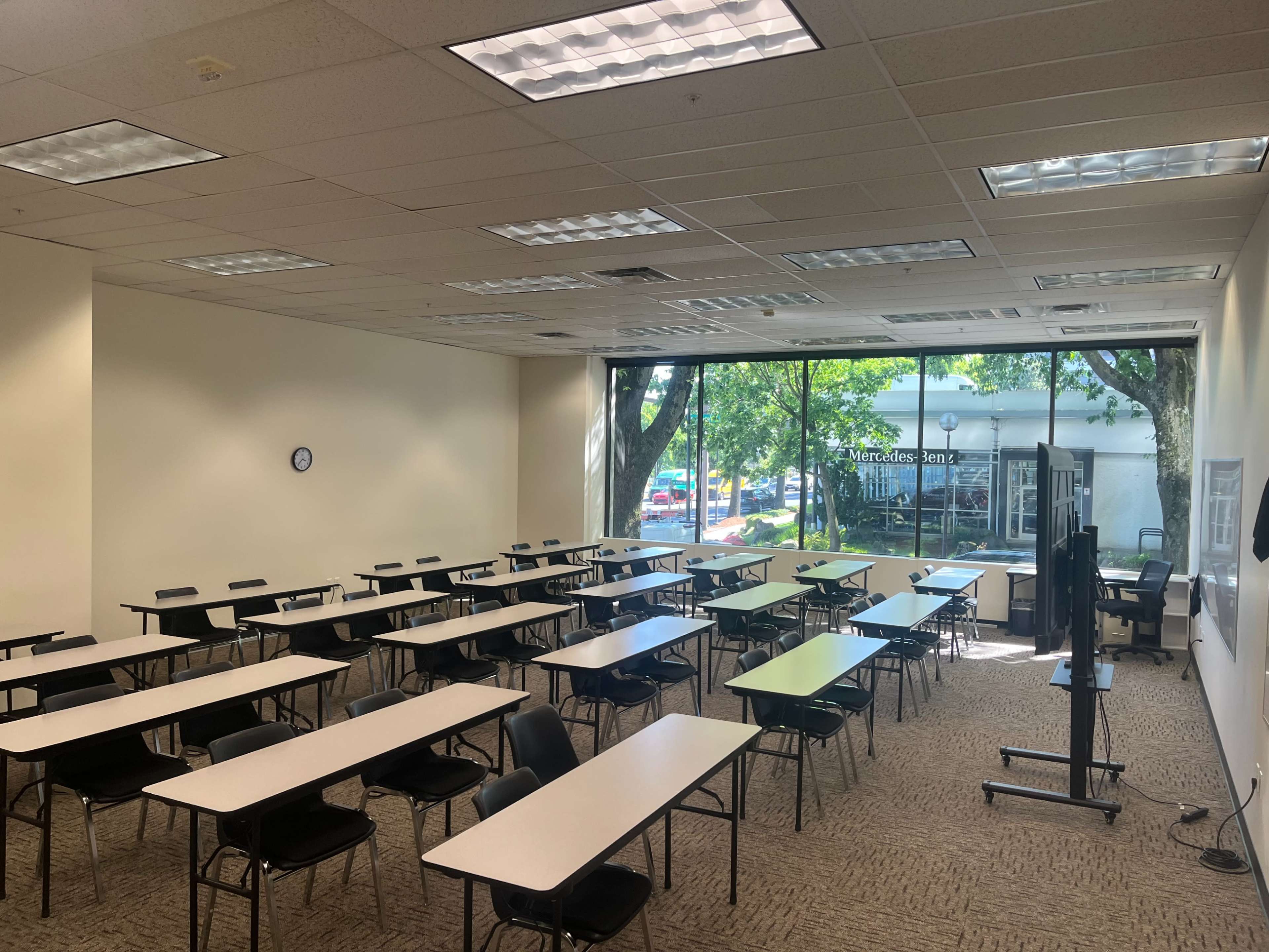 The image shows a spacious classroom arranged with tables and chairs, featuring large windows that offer a view of the outside.