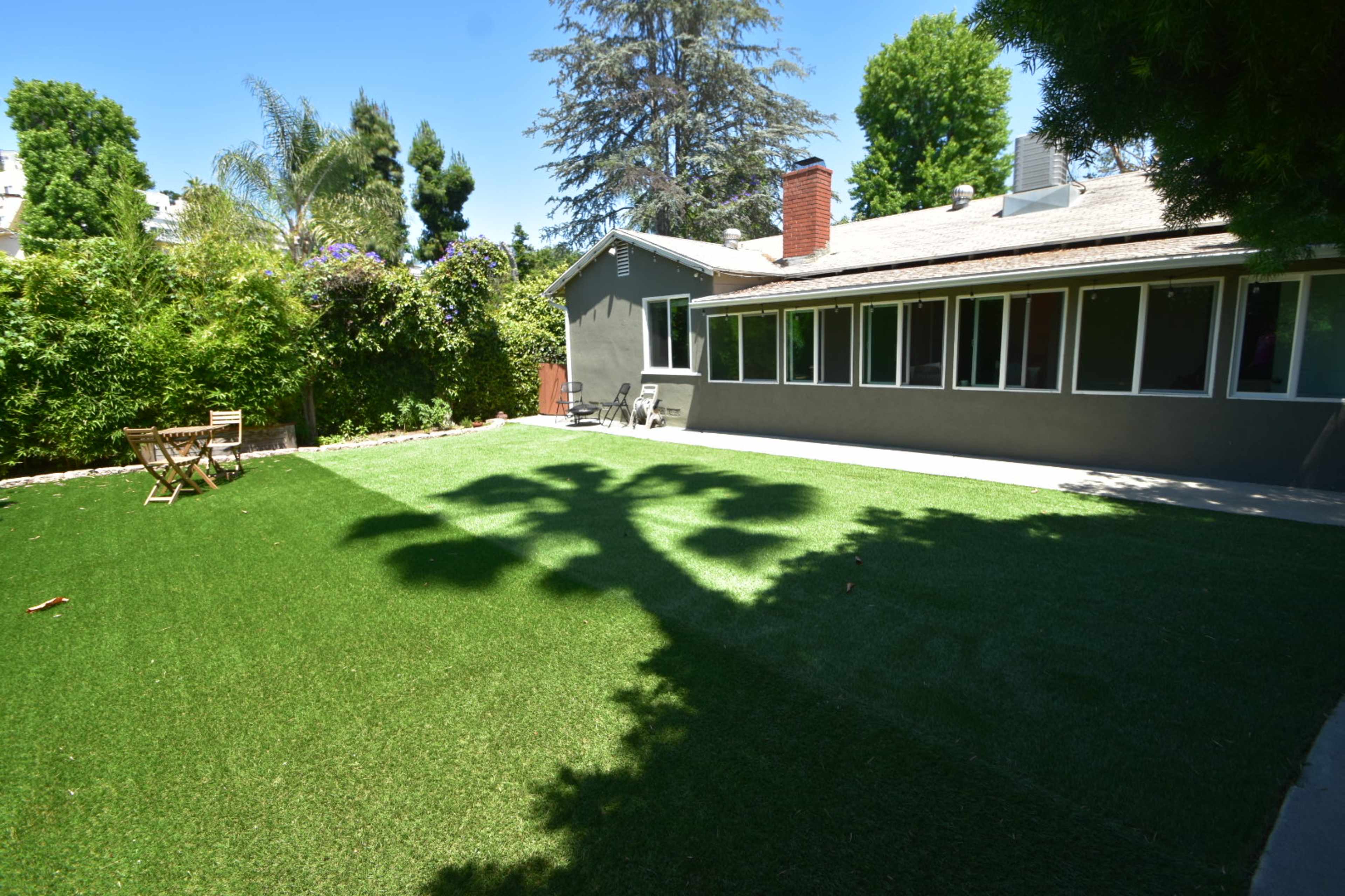 A backyard with artificial grass, featuring a patio area and a few chairs, surrounded by greenery and trees.
