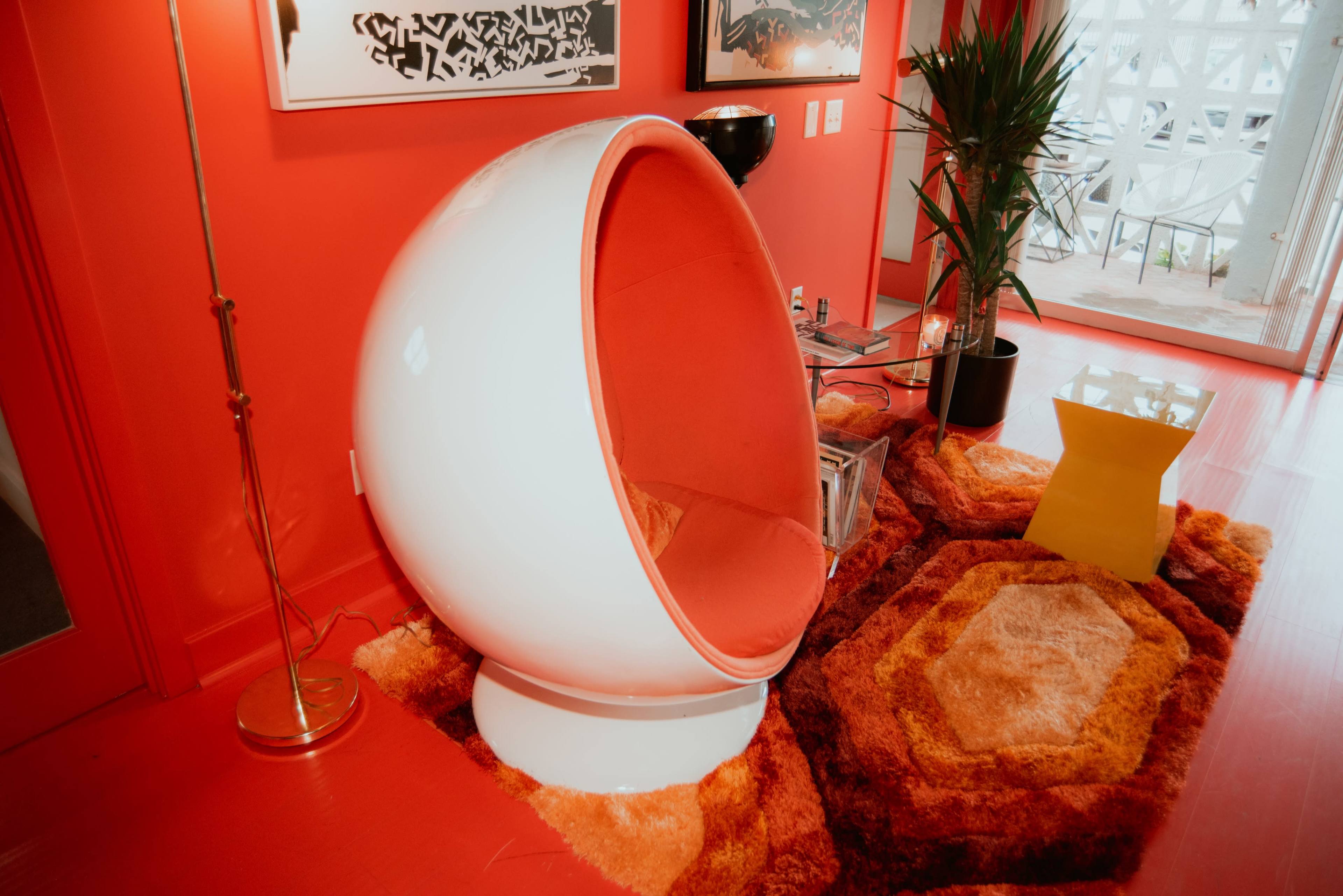 A vibrant red room featuring a white egg-shaped chair with orange upholstery, a colorful shag rug, and a tall plant beside a glass table.