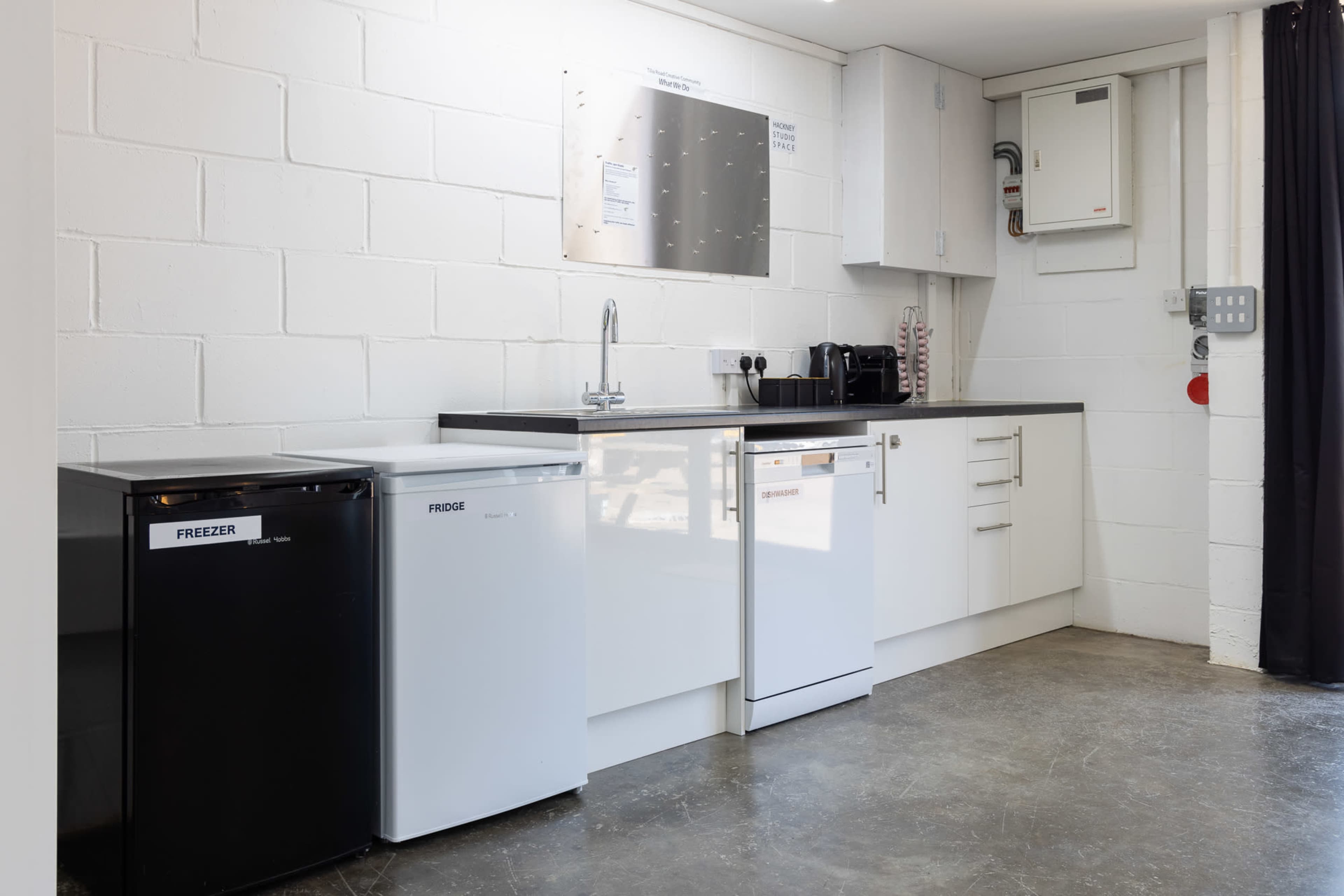 A simple kitchen area with a black freezer, a white refrigerator, and a white dishwasher along a wall with cabinets and a sink.