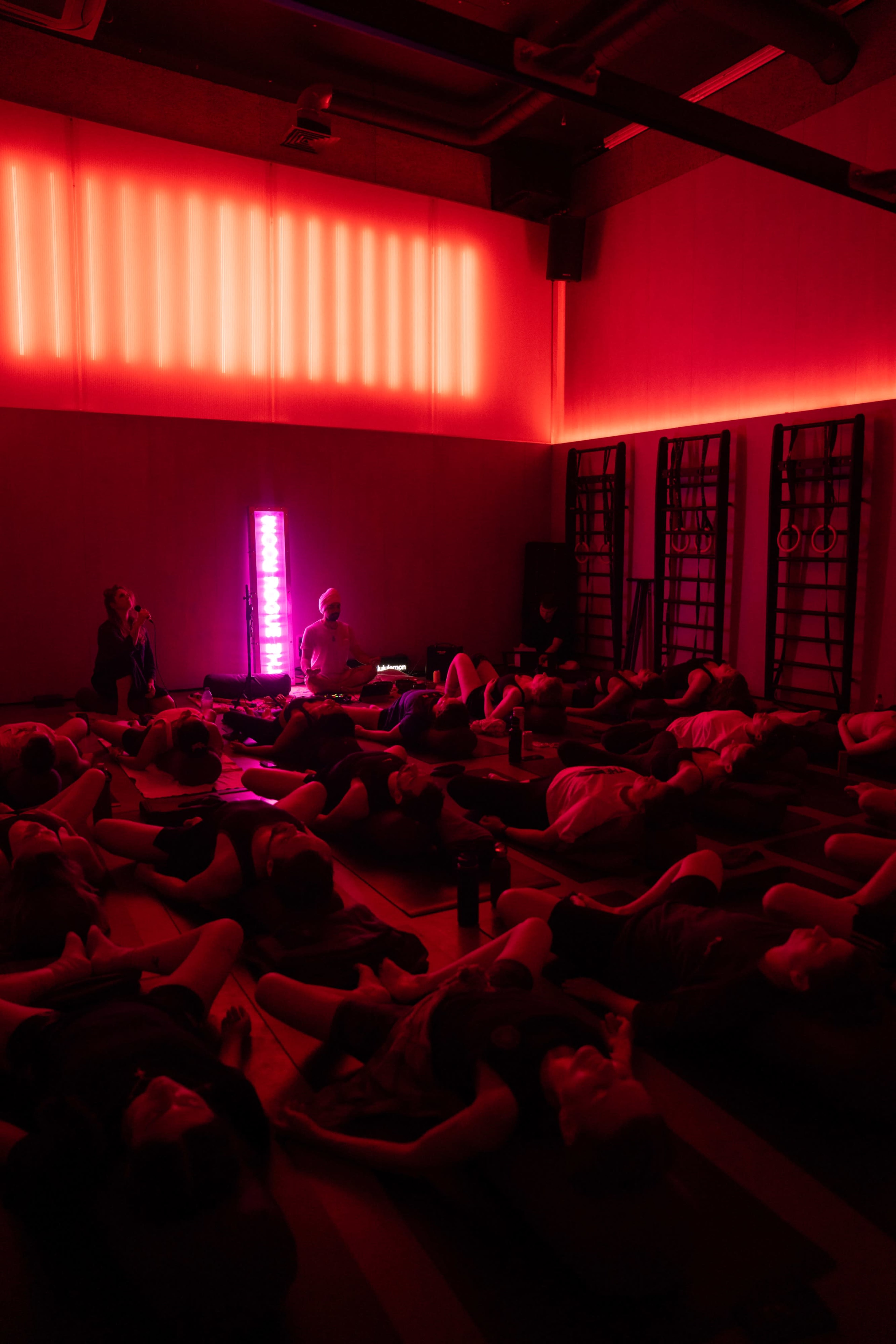 A group of people lie on mats in a dimly lit room with red lighting, while an instructor sits in the center.
