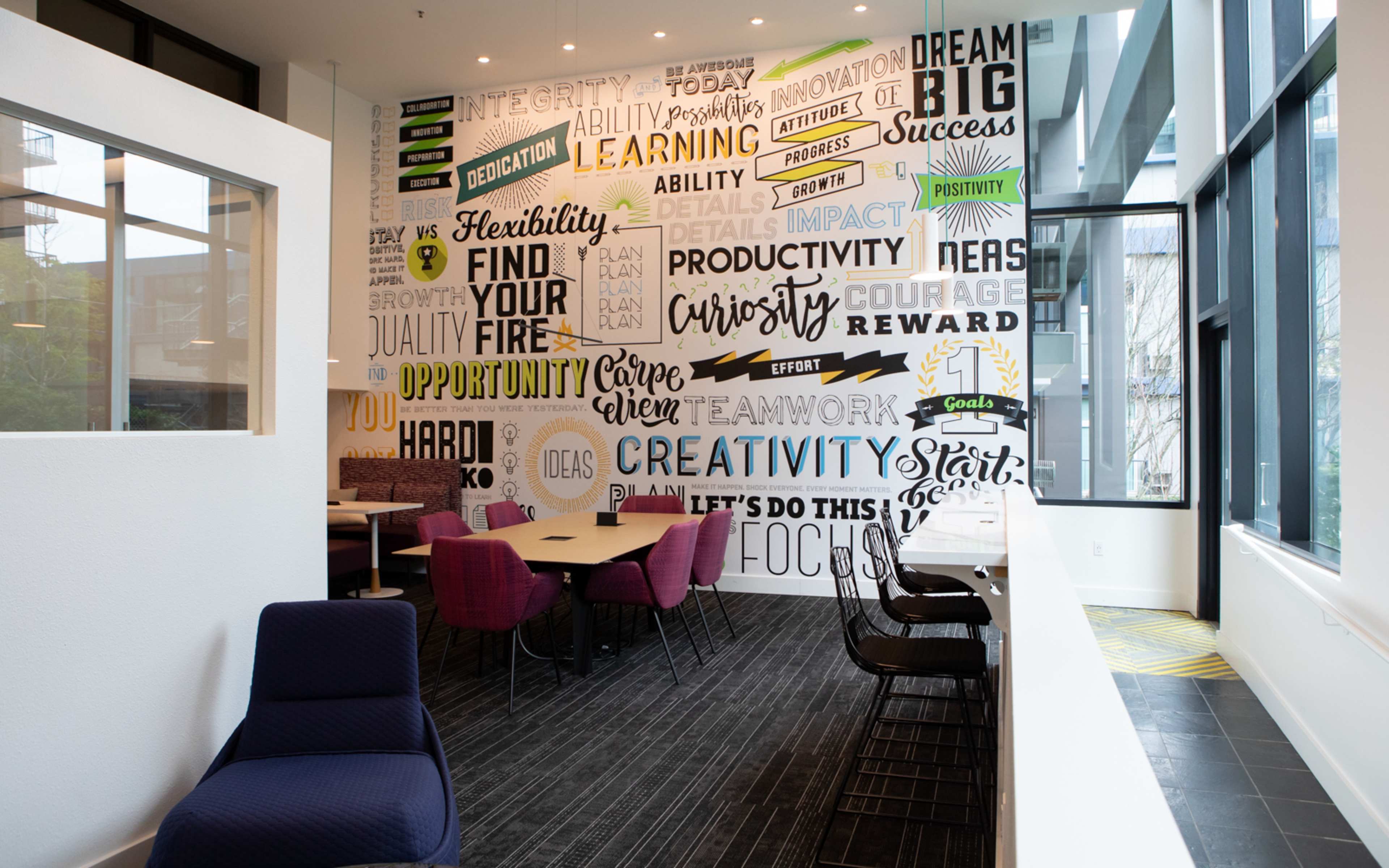 A modern workspace features colorful wall graphics with motivational words and phrases, communal tables, and seating areas.