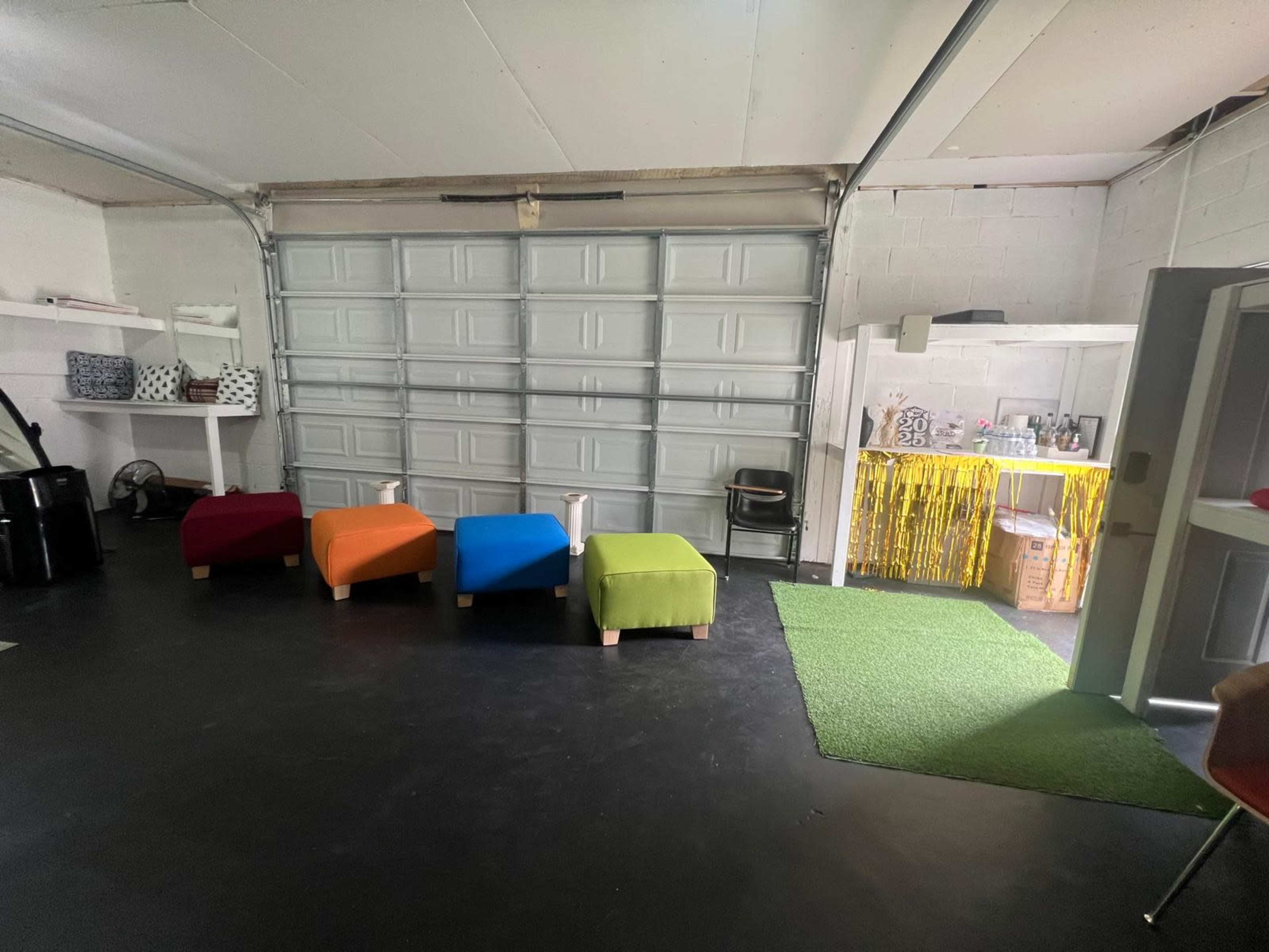The image shows a garage space with five colorful ottomans arranged in a line, a kitchenette area in the background, and a green carpet.