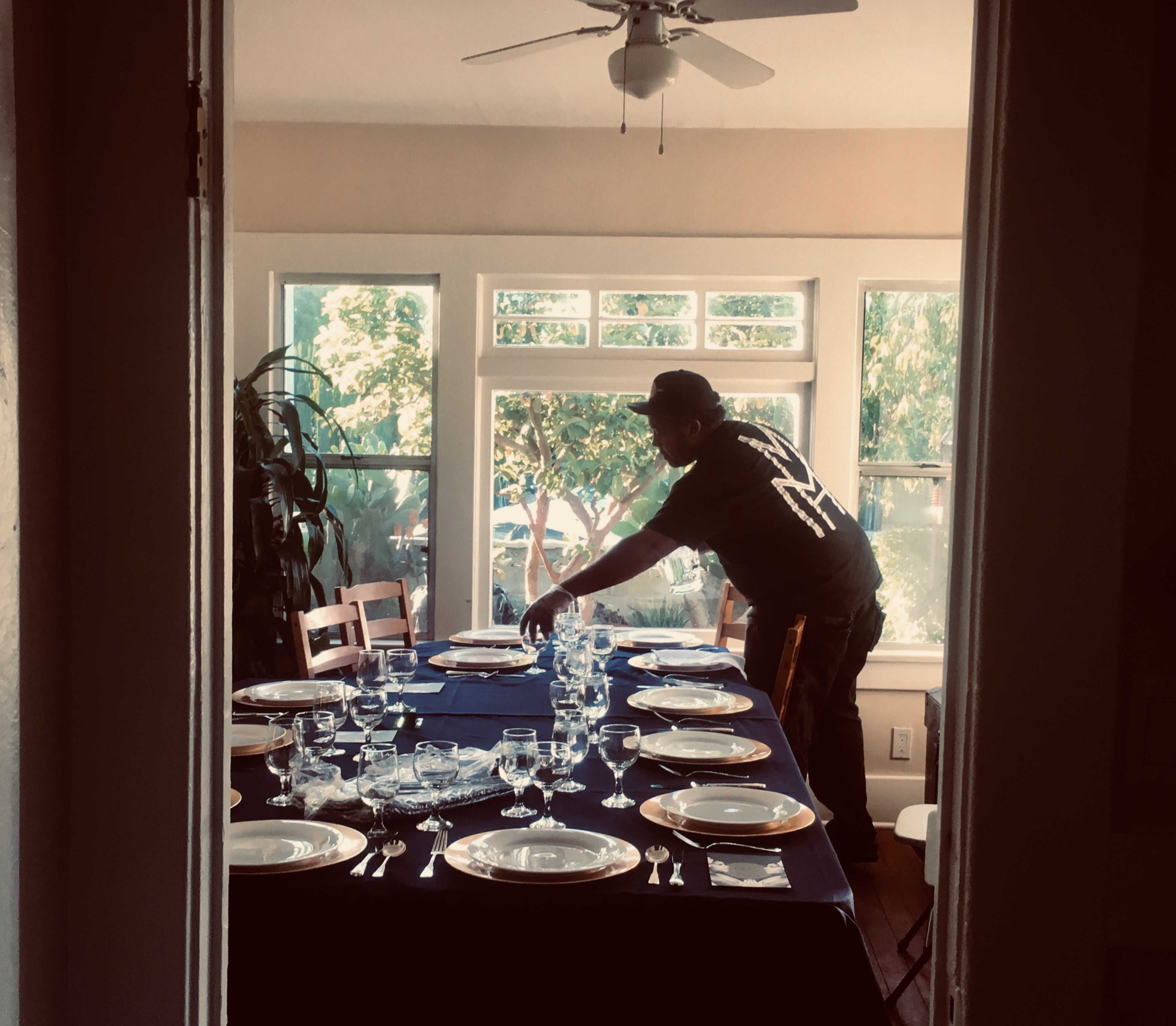 A person sets the dining table in a well-lit room with large windows.