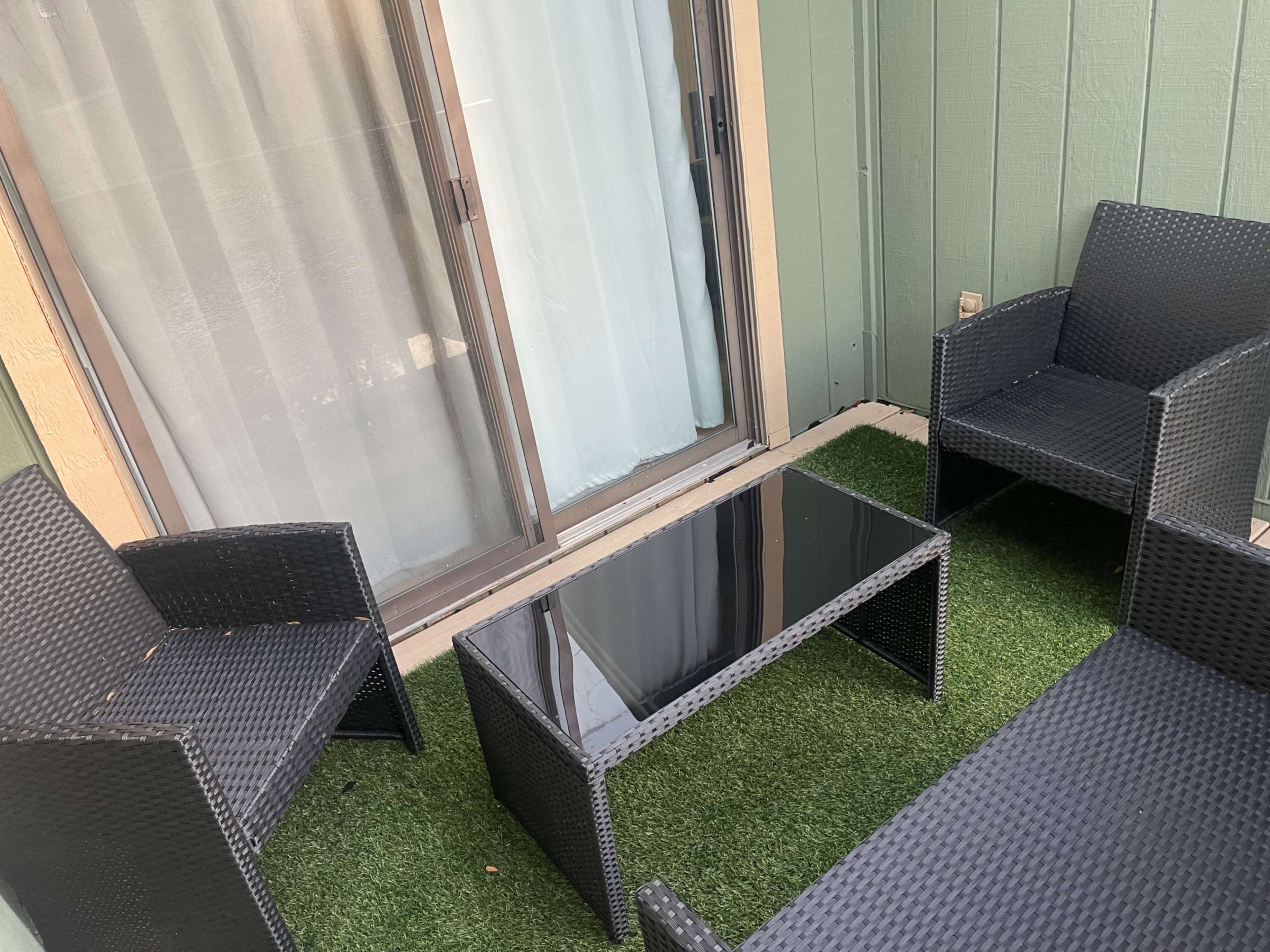 A small outdoor seating area features four black wicker chairs arranged around a glass-top table on green artificial grass.