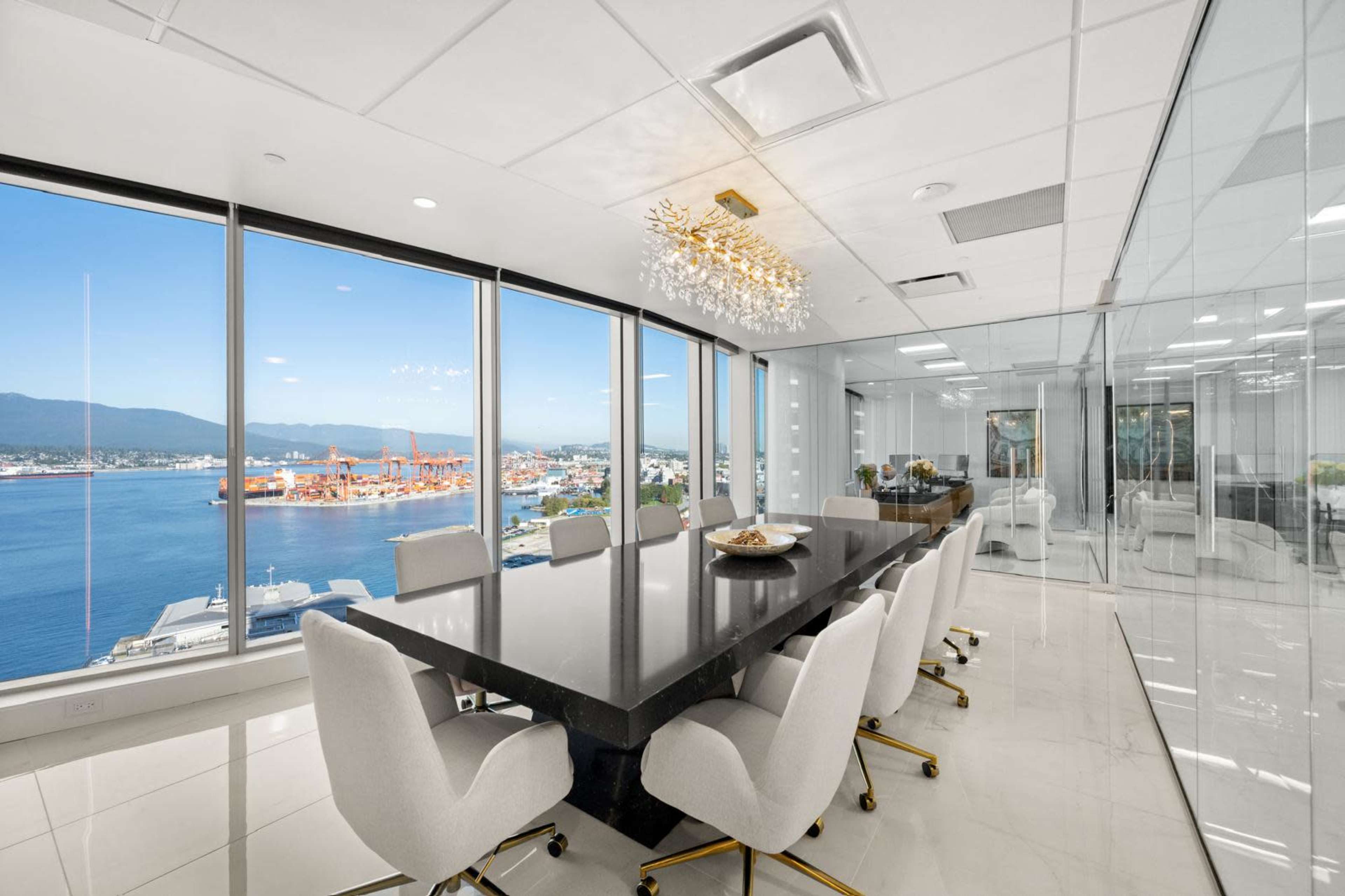 A modern conference room featuring a large black table surrounded by white upholstered chairs, with floor-to-ceiling windows overlooking a harbor and cityscape.