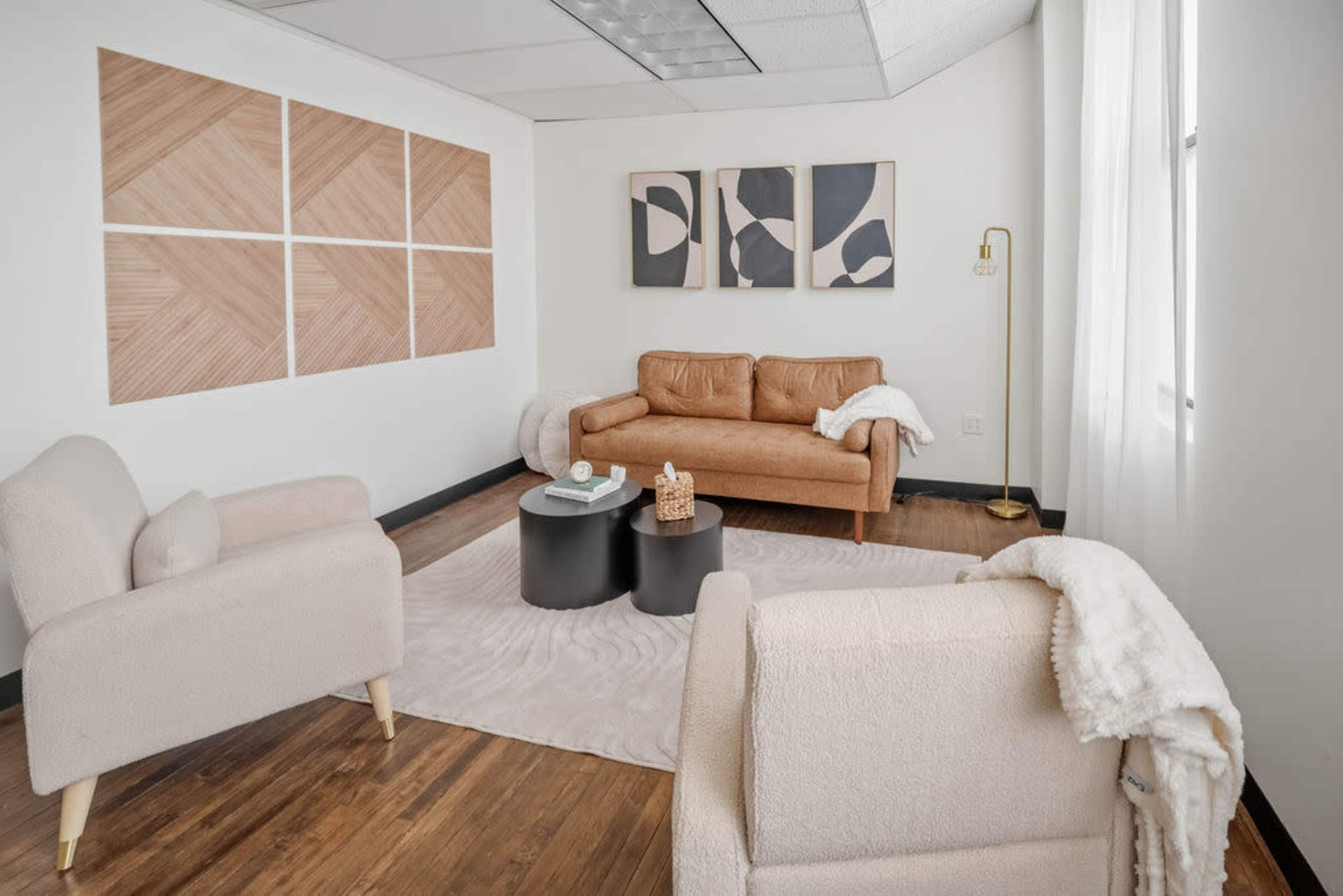 The image depicts a modern lounge area featuring a tan leather sofa, two light-colored armchairs, a coffee table, and abstract wall art.
