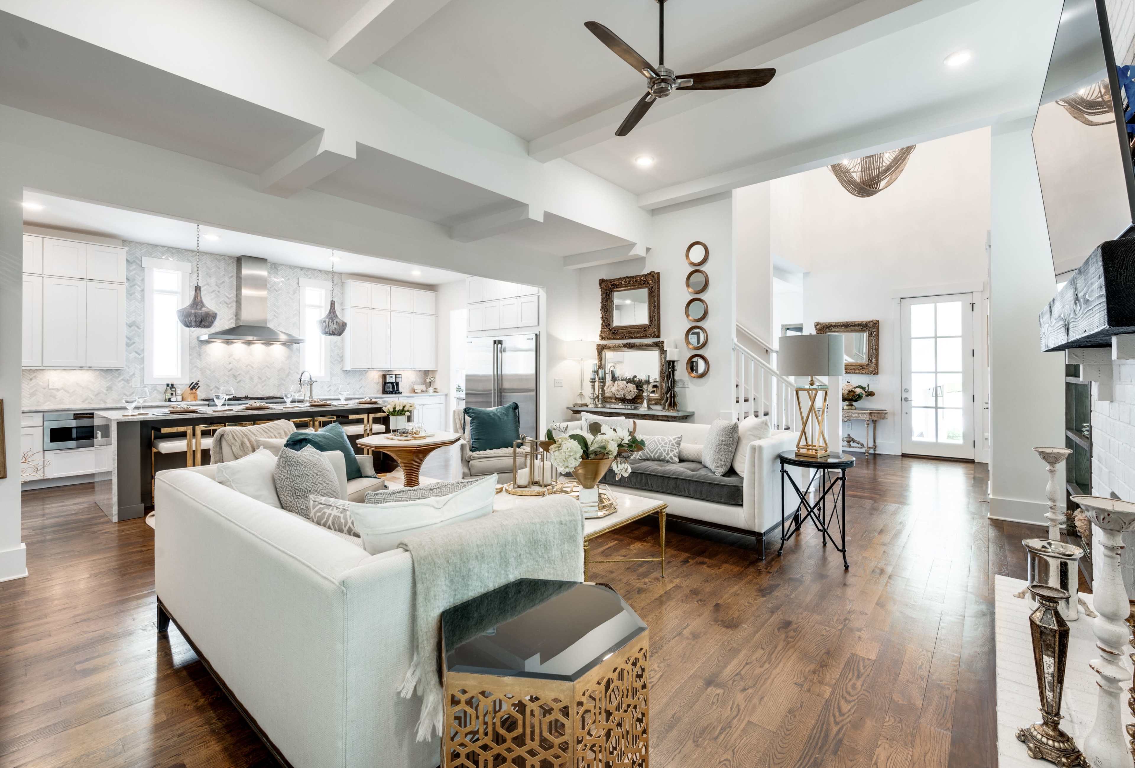 The image shows a modern living room featuring a large sectional sofa, a coffee table, and a kitchen area with a central island, all set against a backdrop of light-colored walls and wooden flooring.