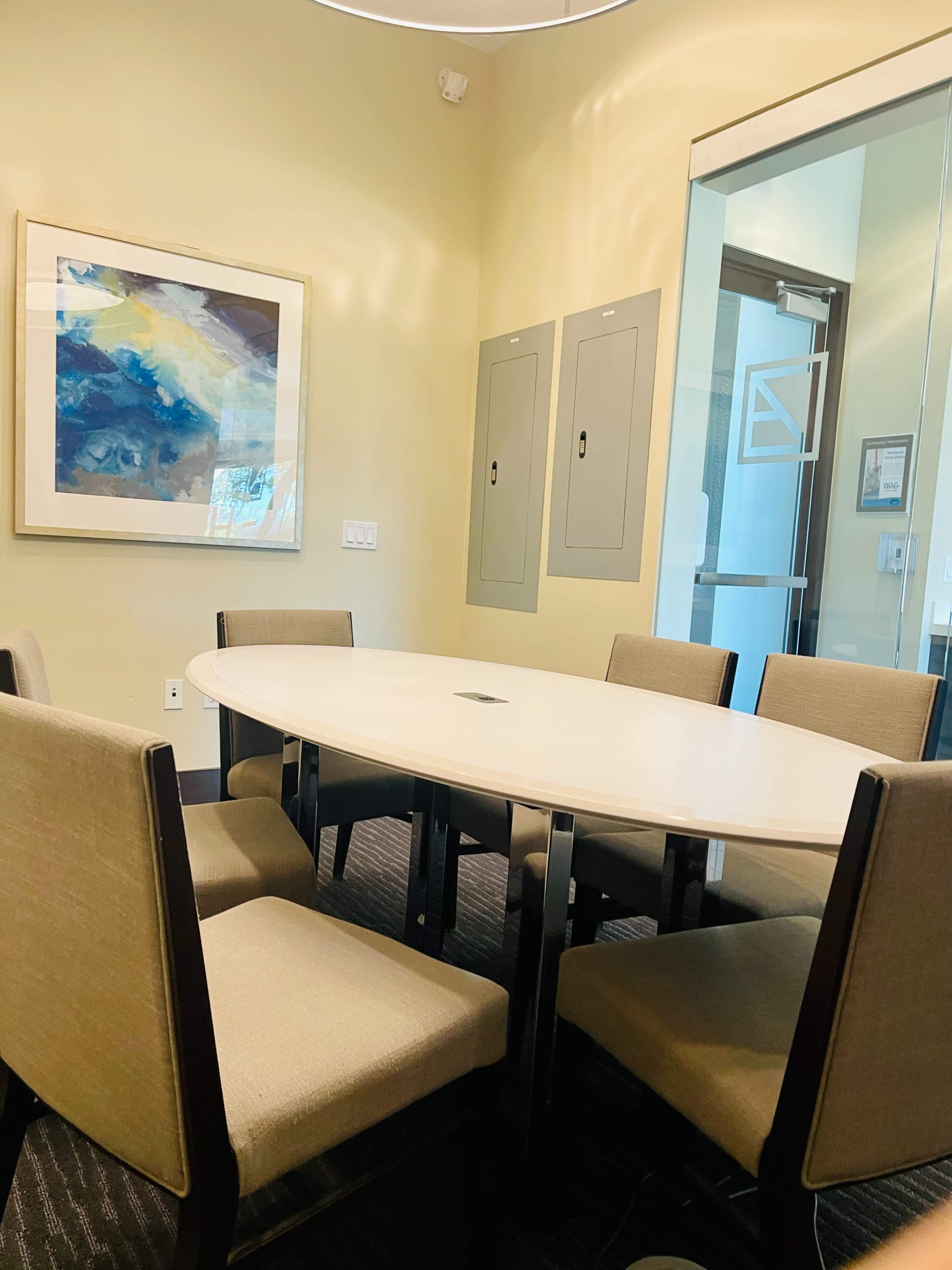 A round table surrounded by six chairs is situated in a small conference room with light-colored walls and a large artwork on one side.