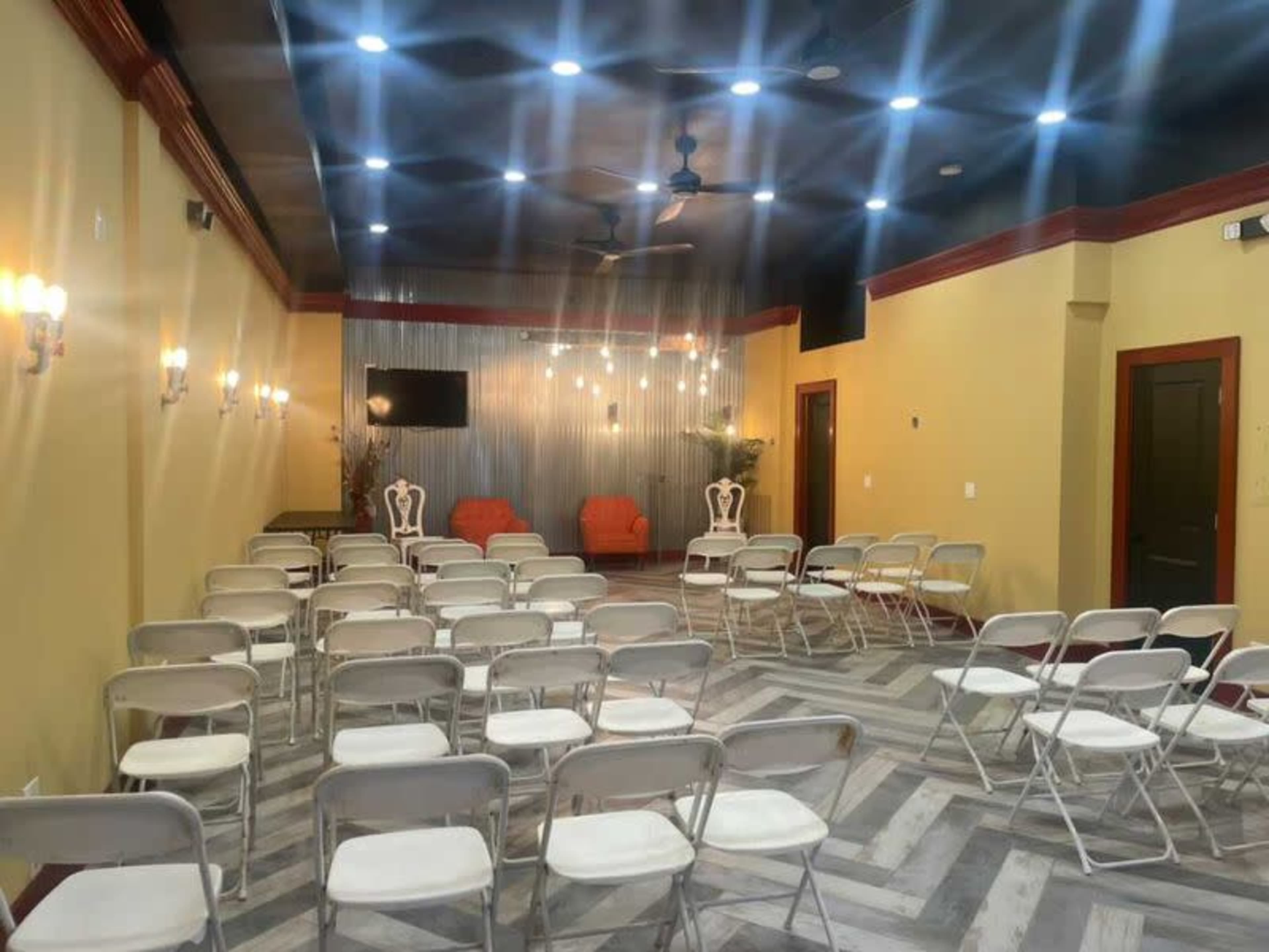 The room features rows of folding chairs facing a decorative wall with two orange chairs and a wall-mounted television, illuminated by overhead lights and sconces.