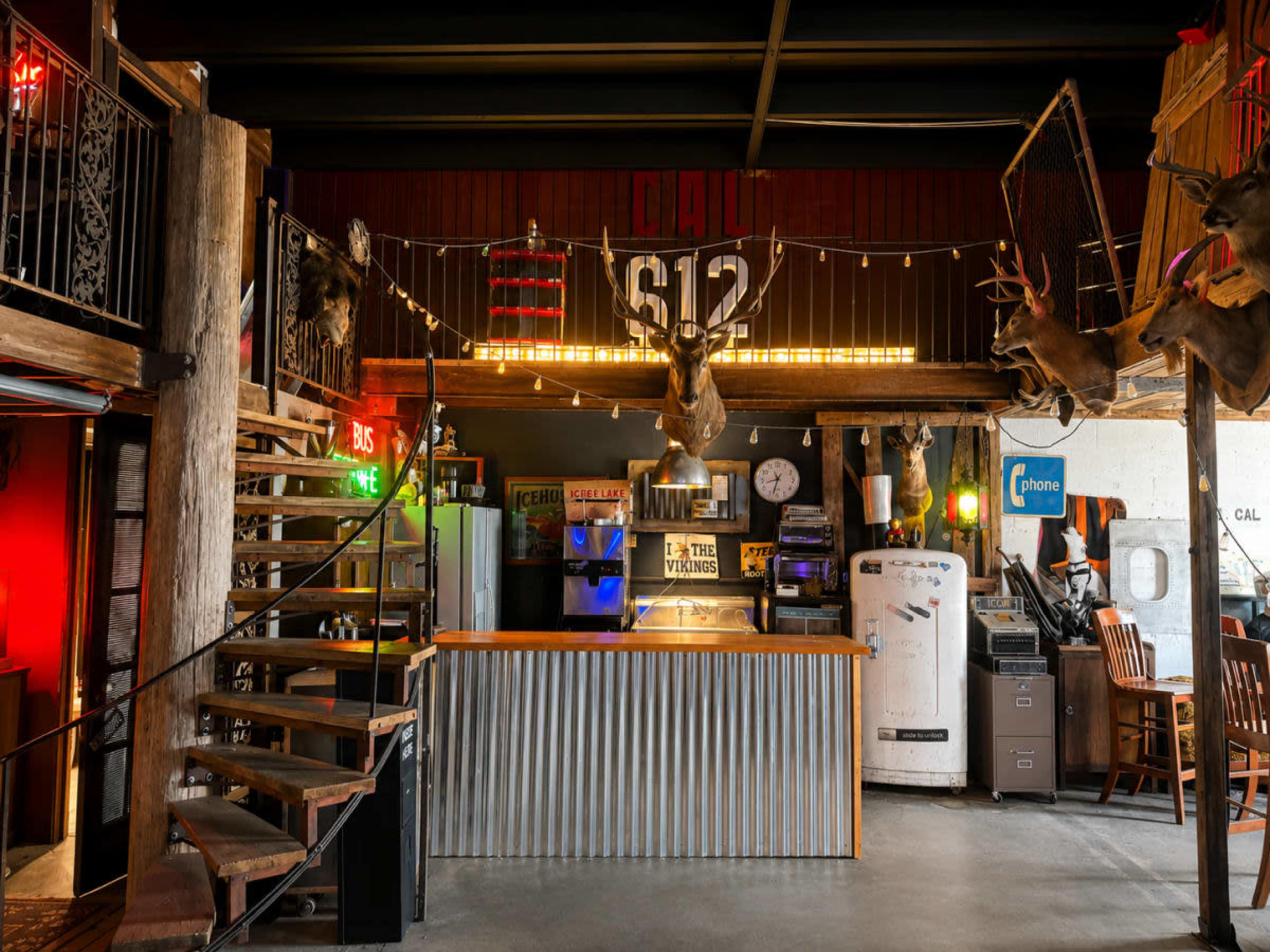 The image shows an interior design of a rustic bar or restaurant with wooden beams, a staircase, various animal mounts on the walls, neon signs, and vintage appliances.