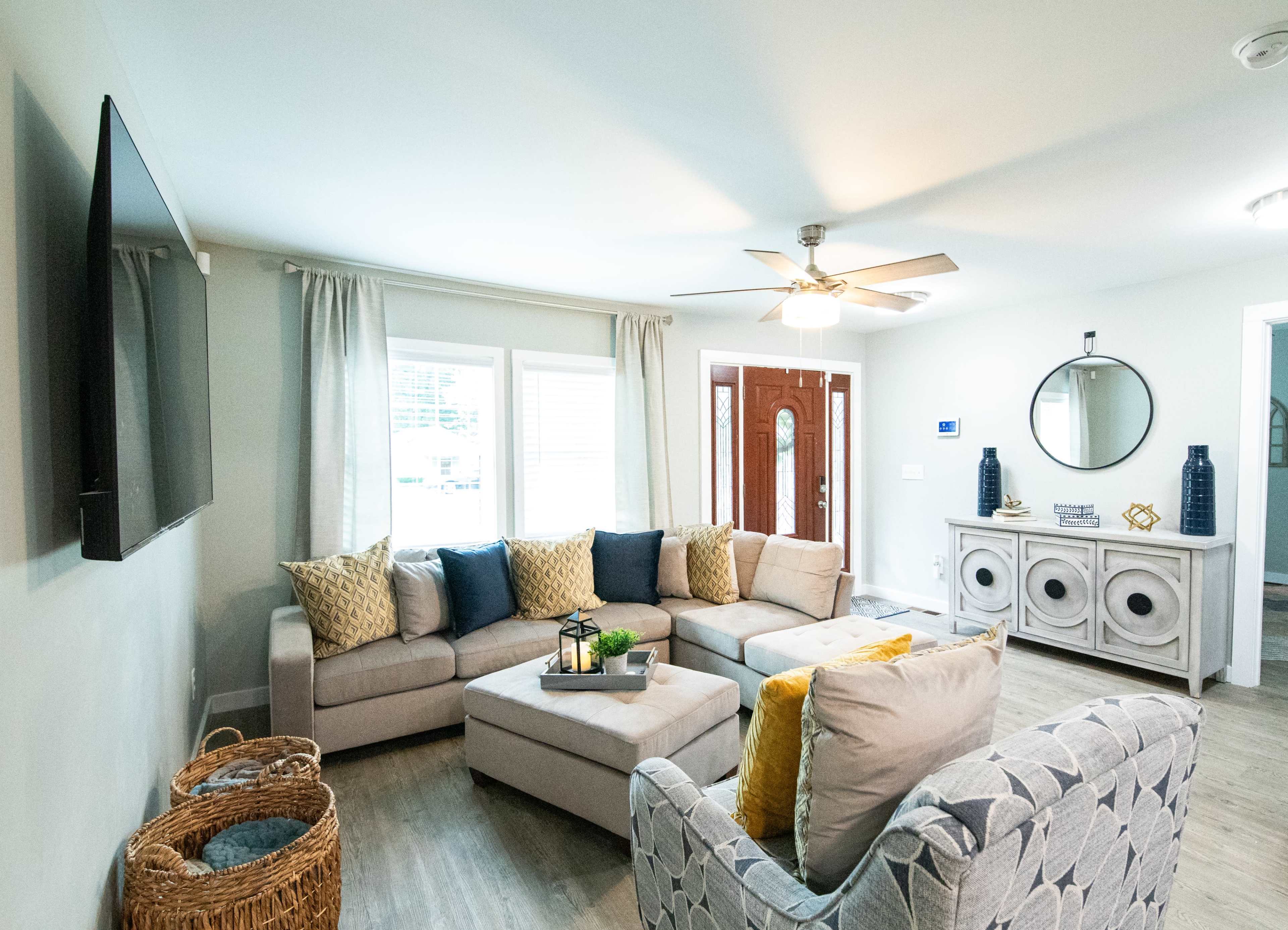 A modern living room features a sectional sofa, a coffee table, a console table, and a decorative mirror, illuminated by natural light from windows.