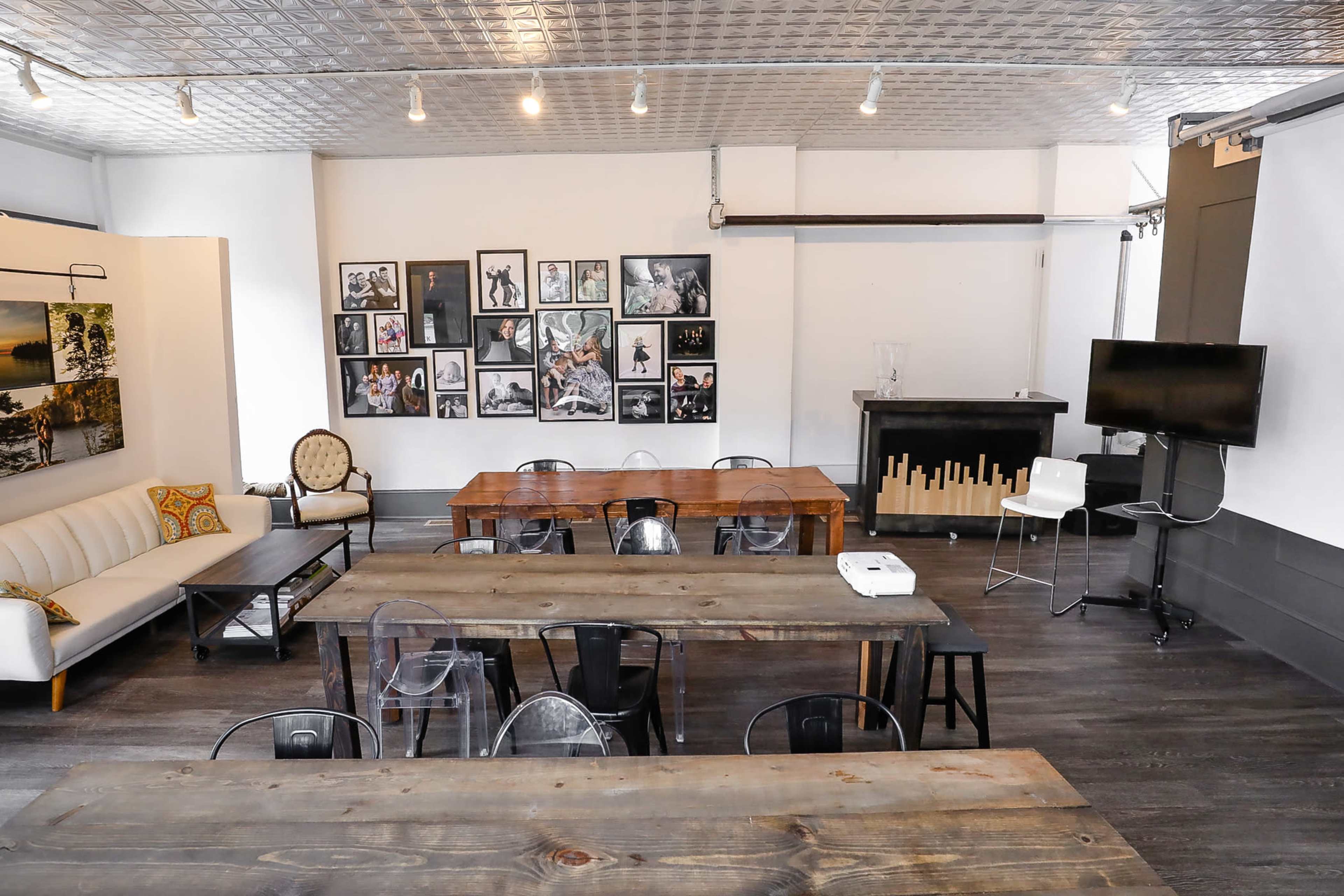 The image shows a spacious, well-lit room featuring wooden tables, a sofa, and walls adorned with framed photographs.