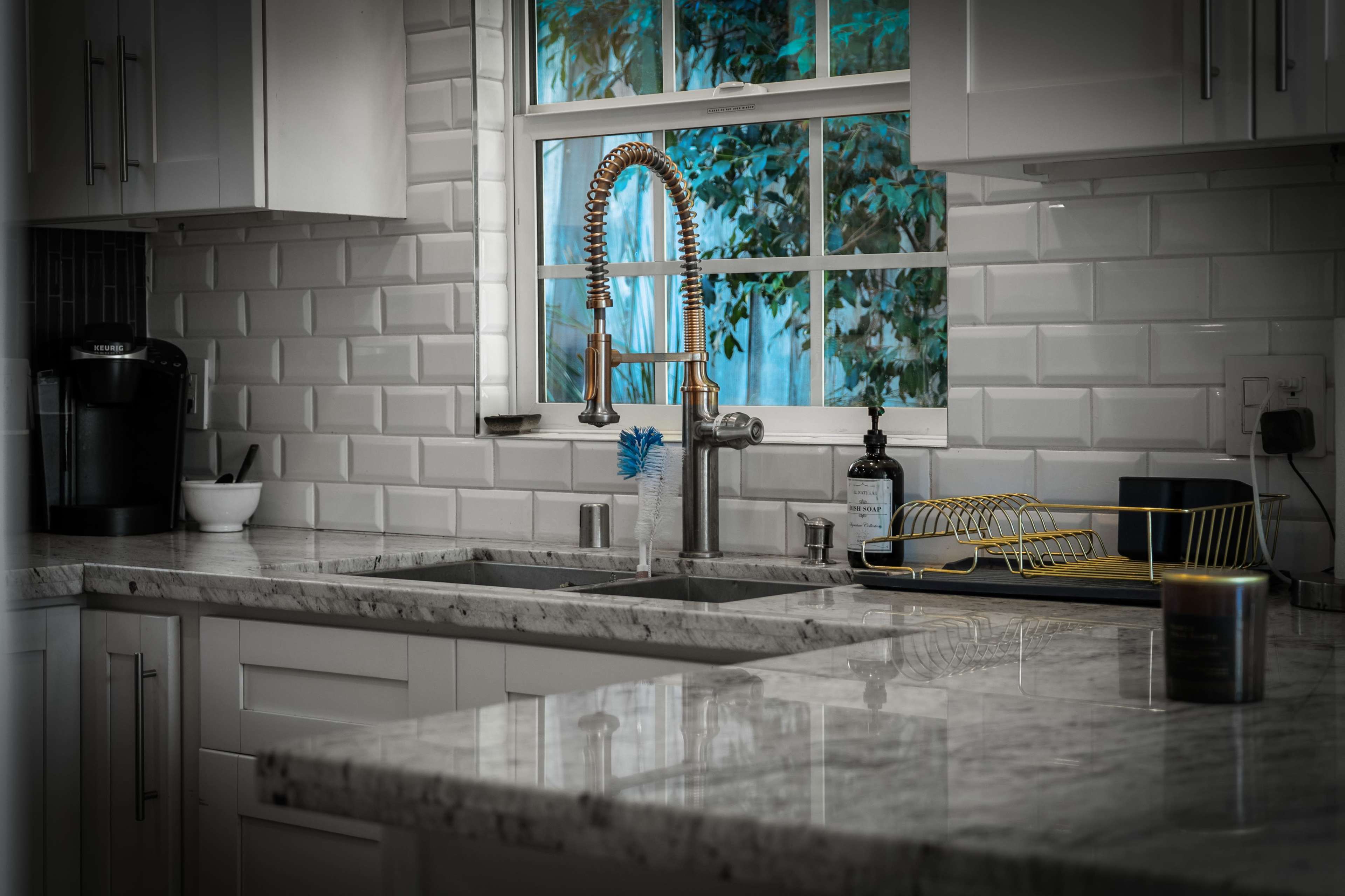 A modern kitchen features a copper faucet, marble countertops, and a window with greenery visible outside.