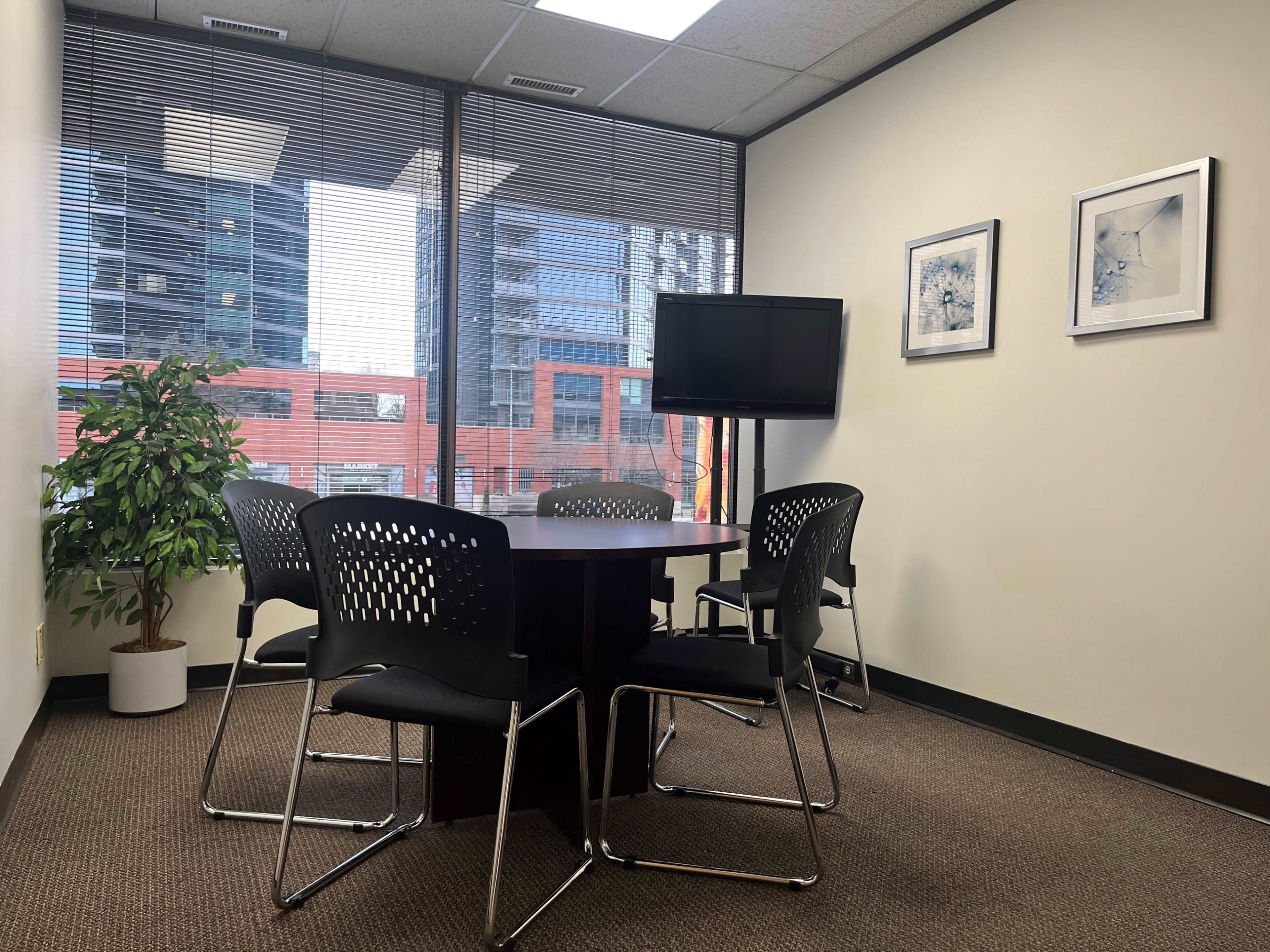 A small meeting room features a round table surrounded by four chairs, with a television mounted on the wall and large windows displaying a cityscape.
