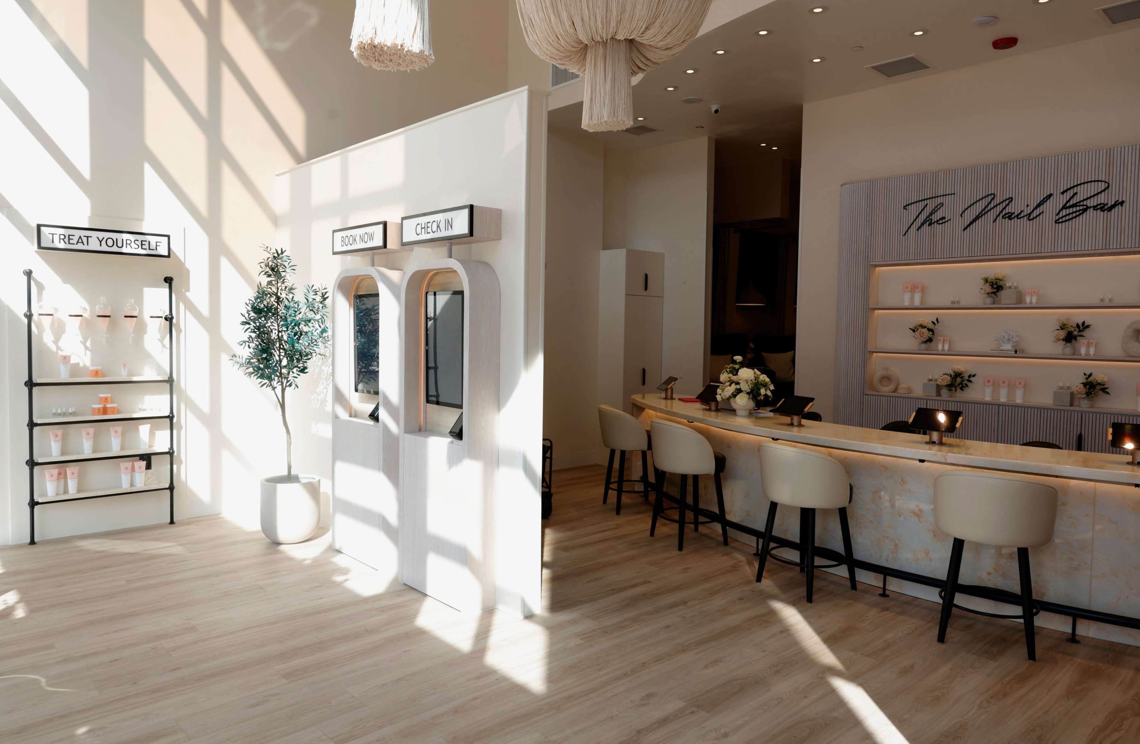The image shows a bright, modern nail salon featuring a reception desk, two check-in mirrors, and a shelf labeled "TREAT YOURSELF" filled with products.