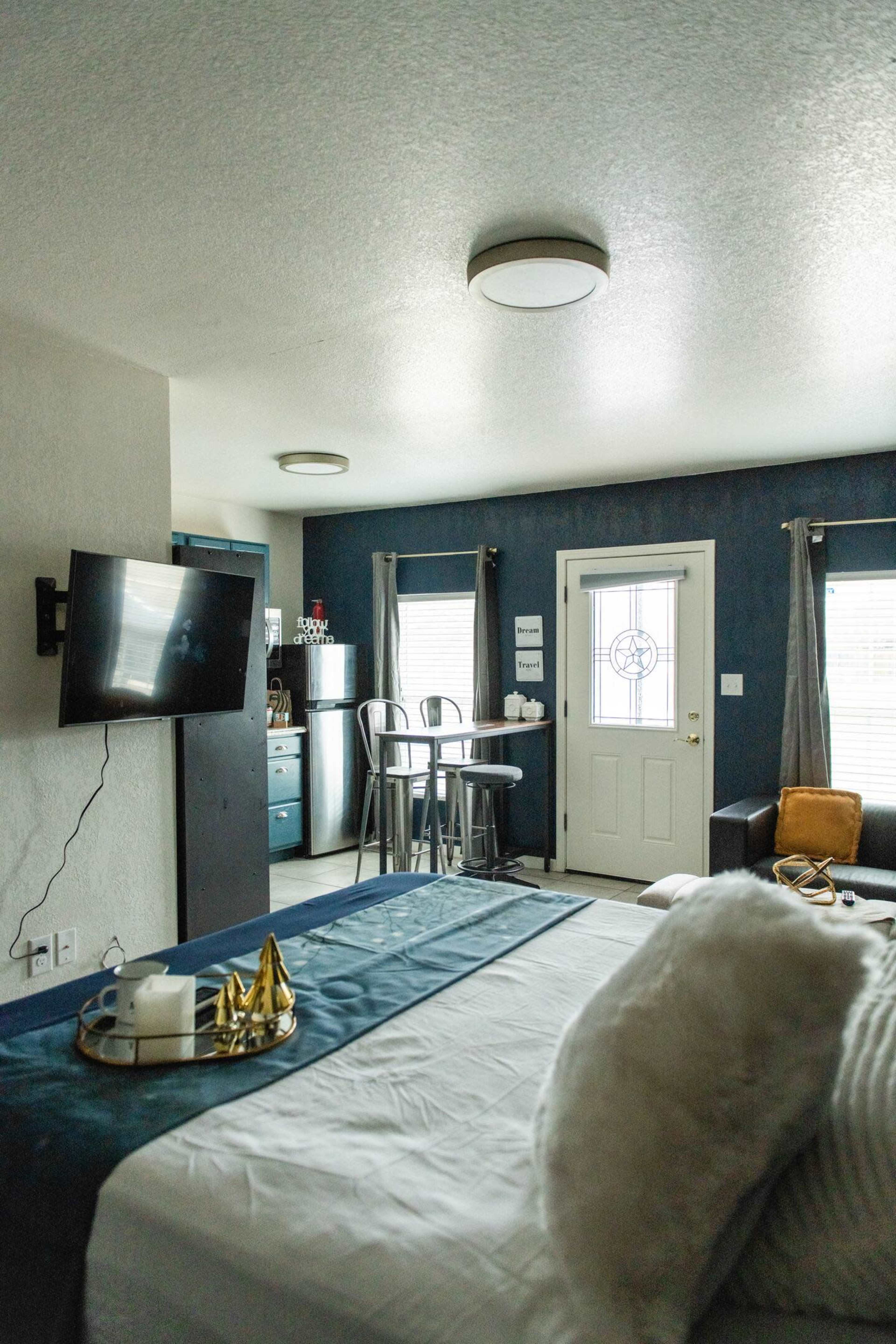 The image shows a cozy studio apartment with a bed, a television mounted on the wall, a small dining area, and a door leading outside.