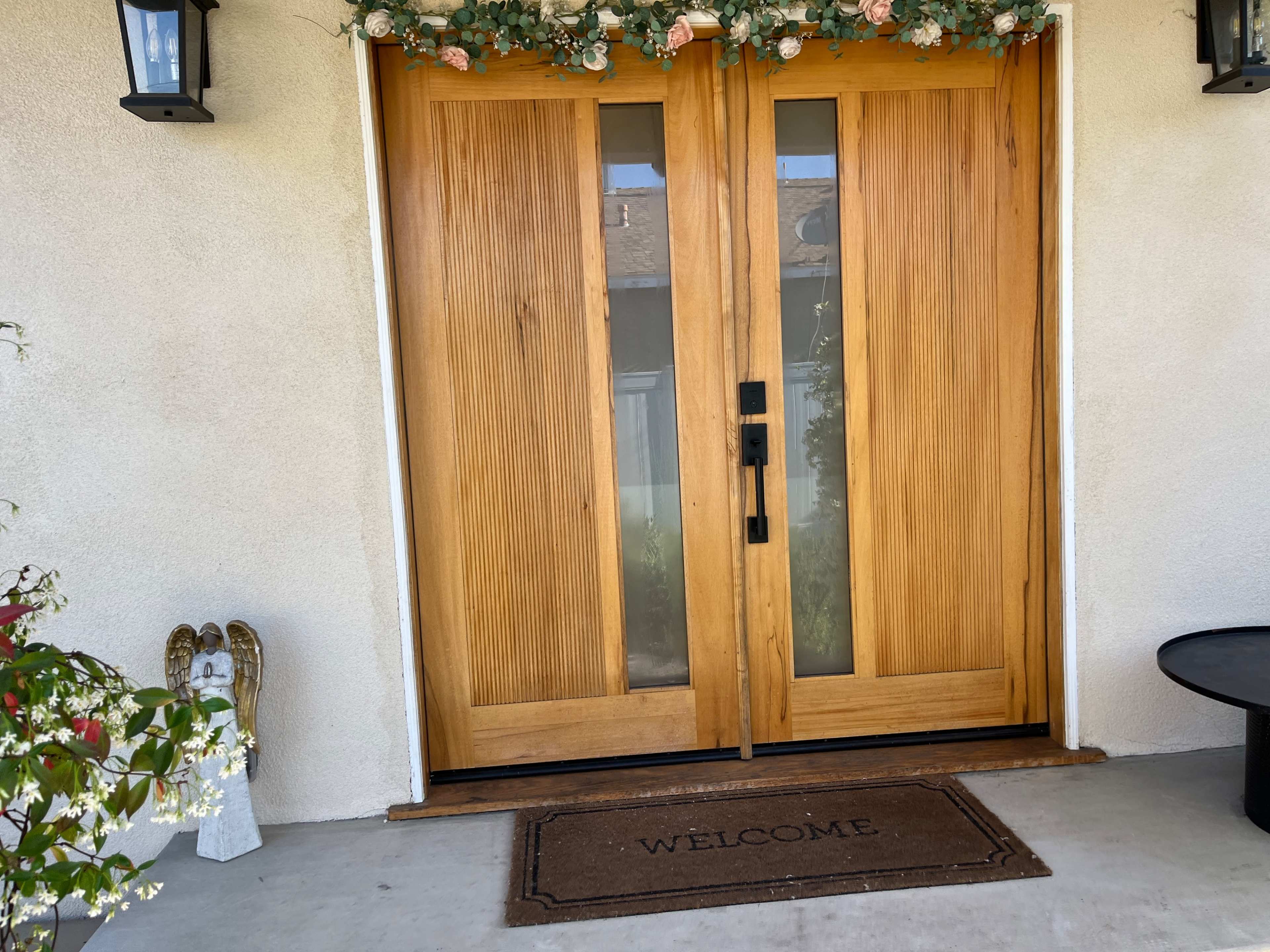 The image shows a pair of wooden double doors with glass panels, flanked by decorative light fixtures and a "WELCOME" doormat at the entrance of a house.