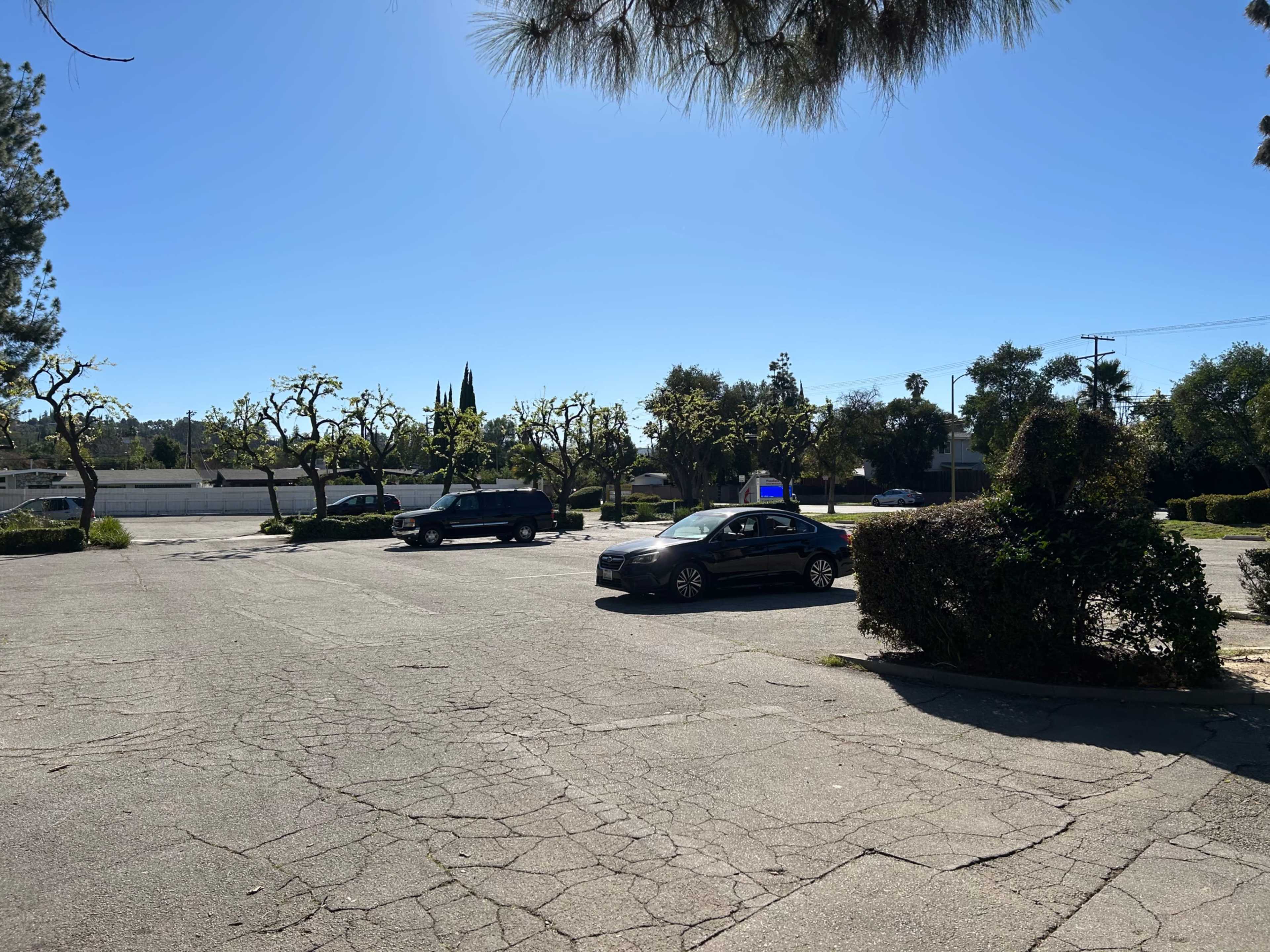 The image shows an empty parking lot under a clear blue sky, with a few parked cars and trees lining the edges.