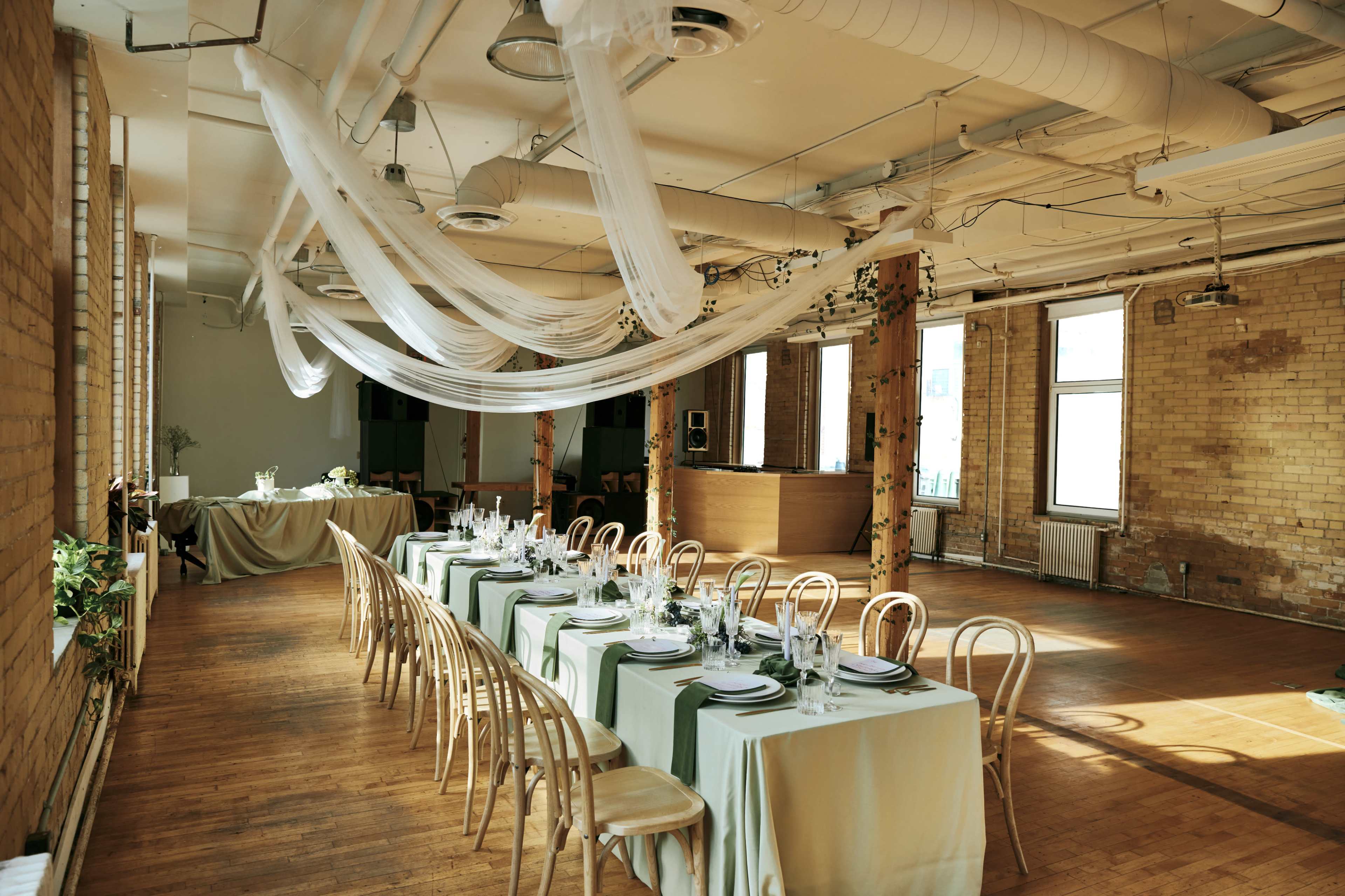 A long banquet table is set with plates and utensils in a spacious, well-lit room featuring exposed brick walls and draped fabric overhead.