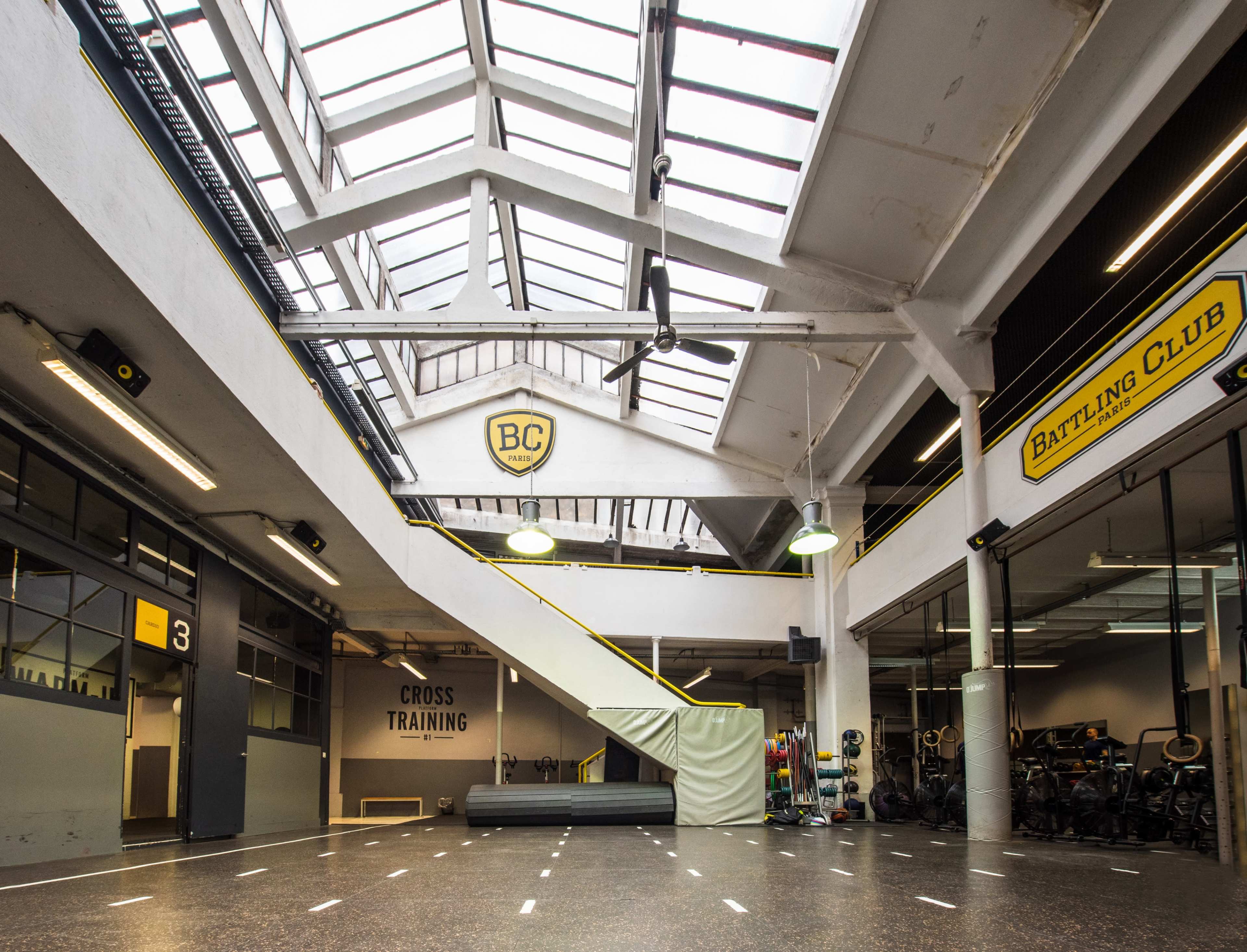 A spacious gym interior featuring large windows along the ceiling, a staircase leading to an upper level, and designated areas for cross training and a banting club.