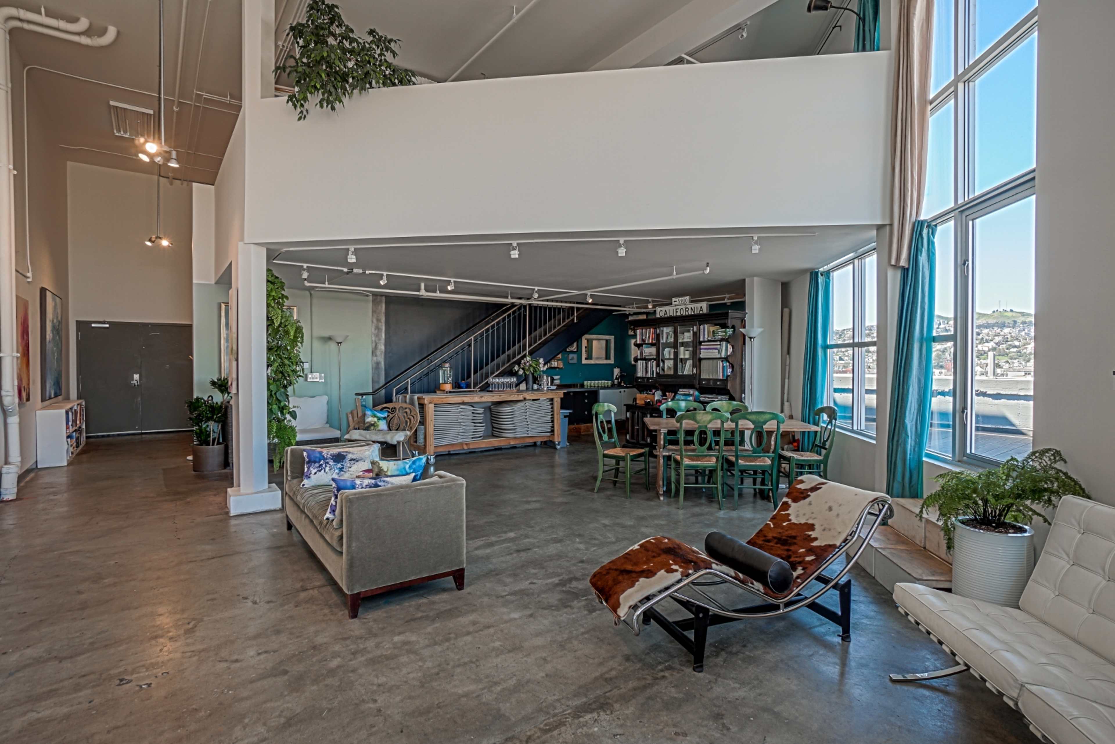A spacious, open-concept room featuring a mix of modern and rustic furniture, large windows, and a staircase leading to an upper level.