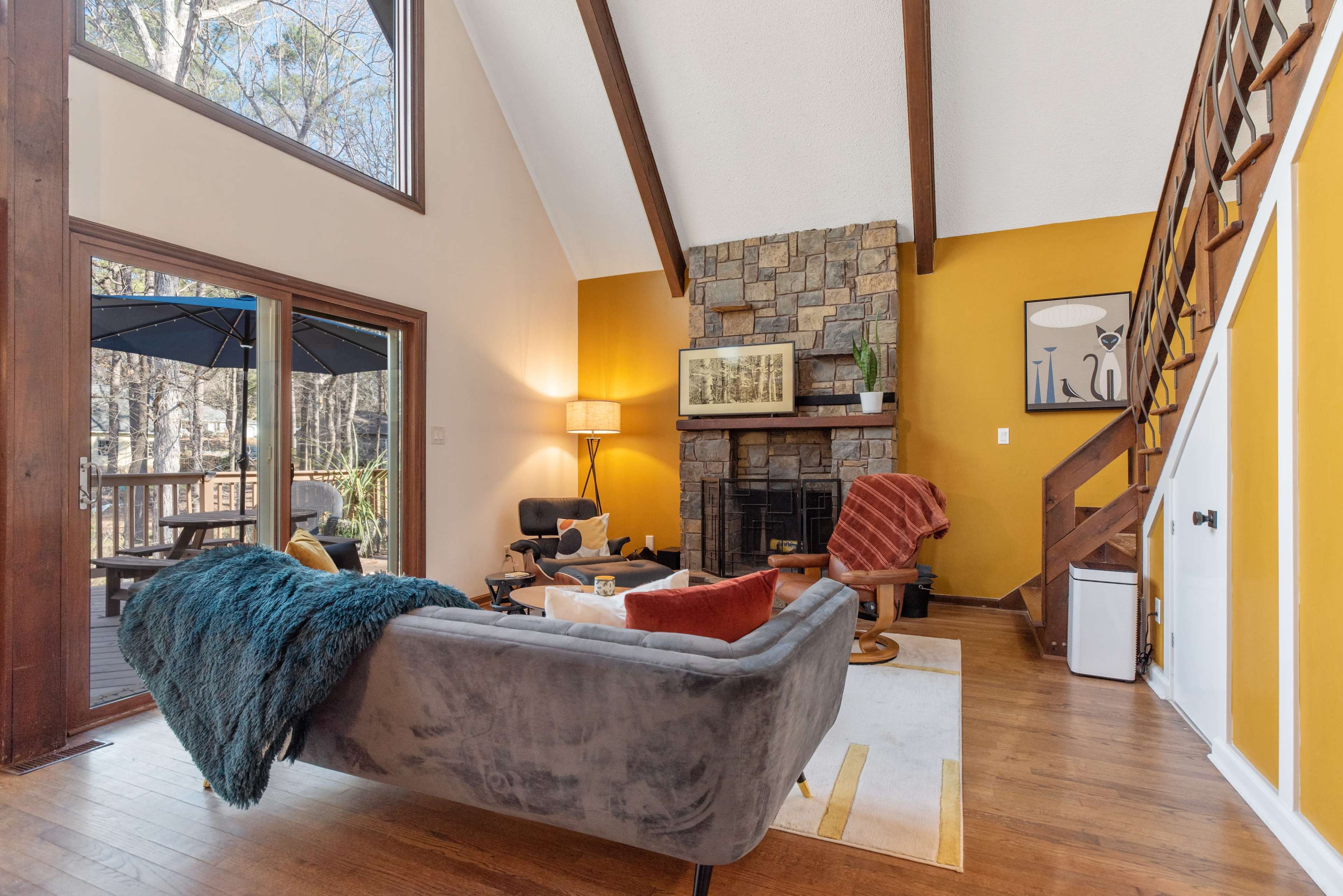 A cozy living room featuring a stone fireplace, large windows, and wooden beams, with a sofa and a small coffee table arranged on a rug.