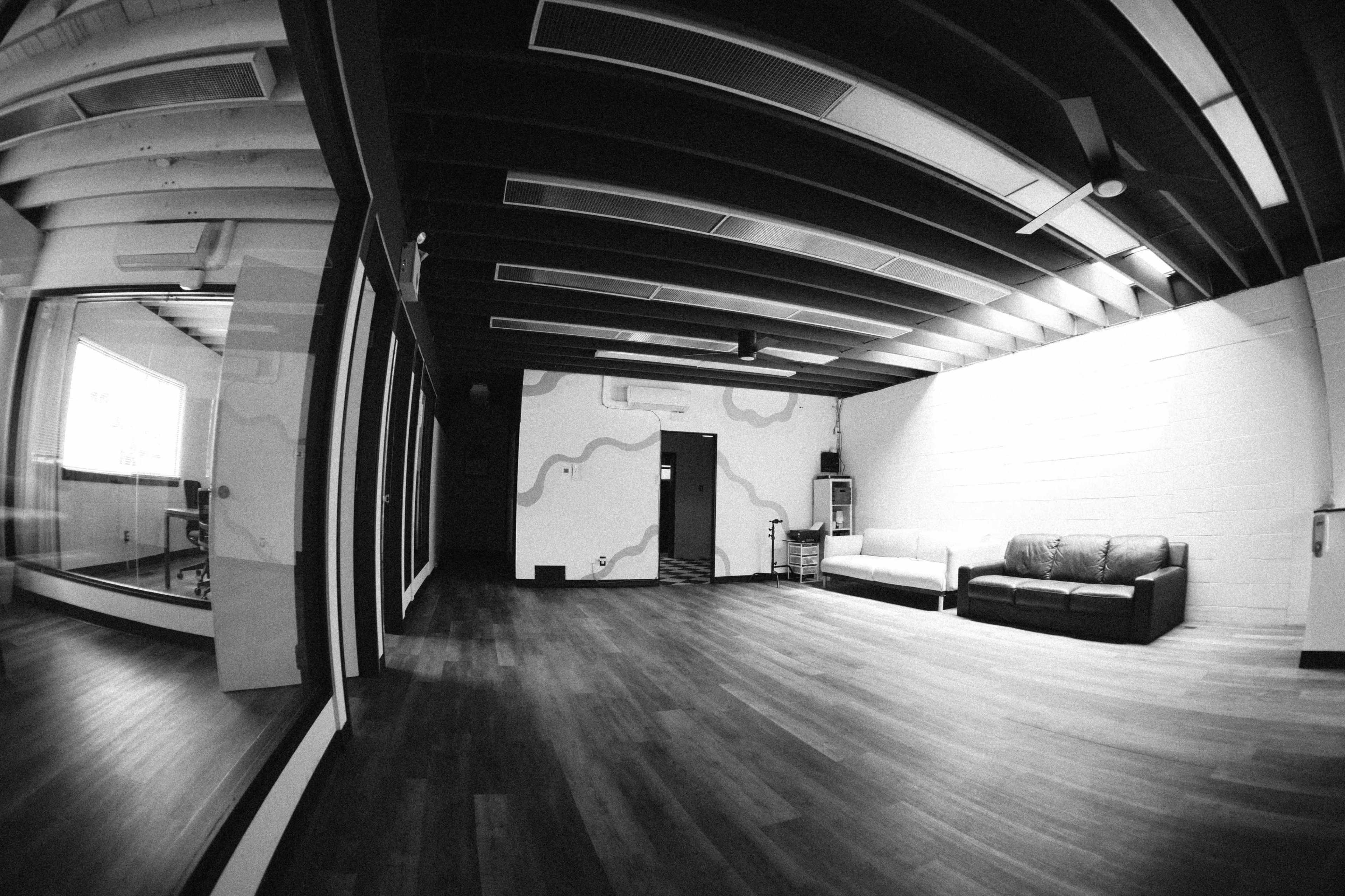 The image shows a spacious, empty room with wooden floors, exposed ceiling beams, and a couch, along with a glass-walled area featuring a chair.